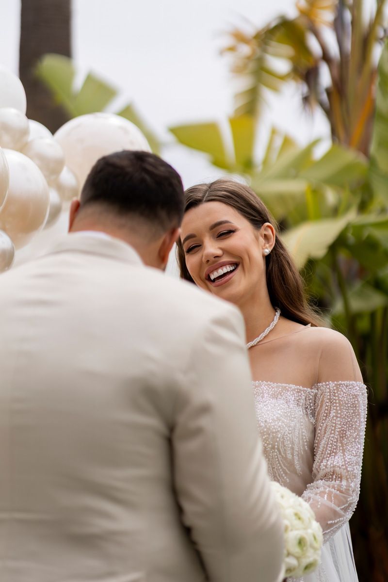 A bride laughs brightly at her groom while standing in front of a neutral balloon arch during an Alicante destination wedding.