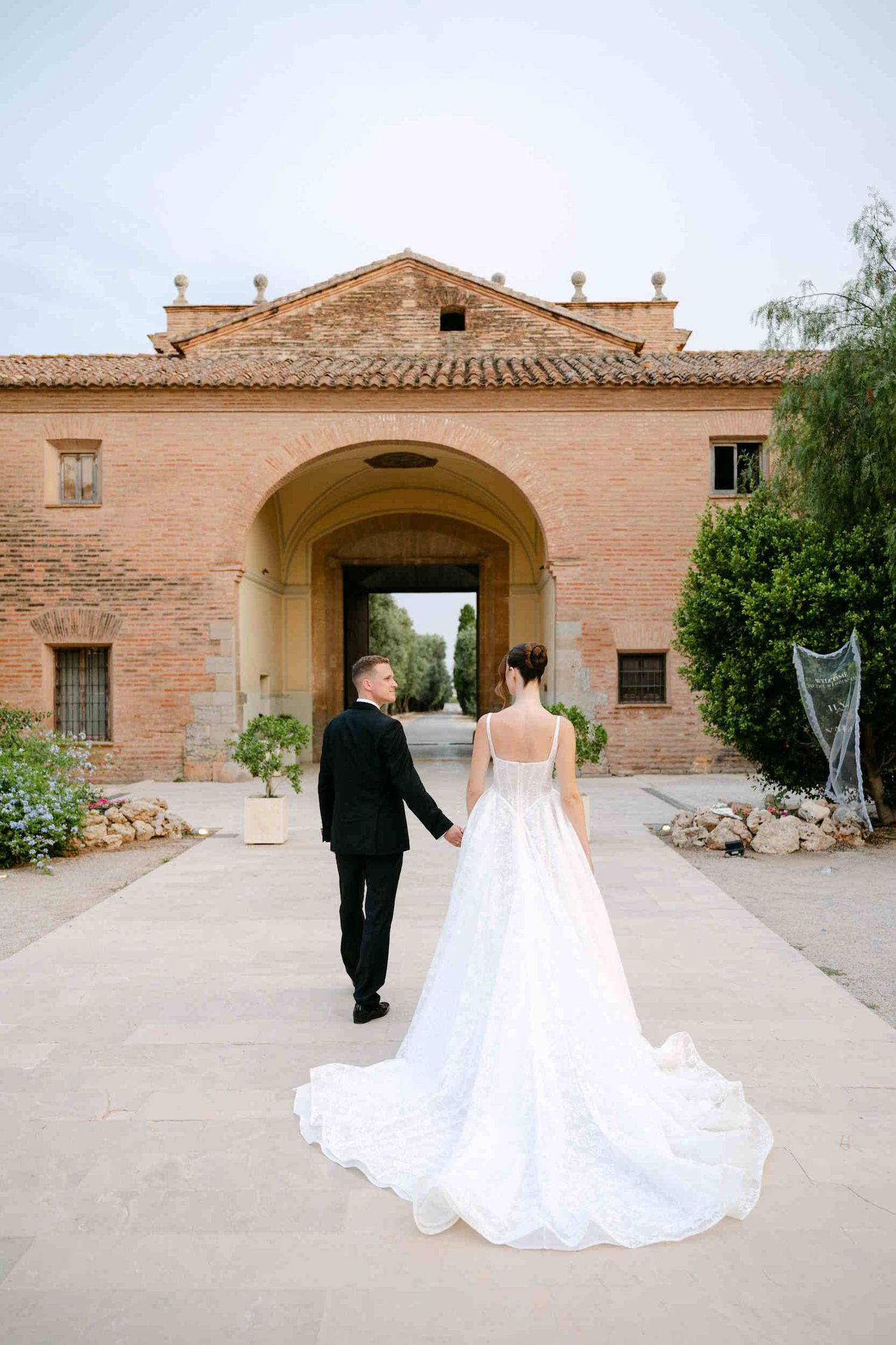 Bride and groom holding hands while walking away across a paved courtyard toward a massive brick archway at the wedding venue.