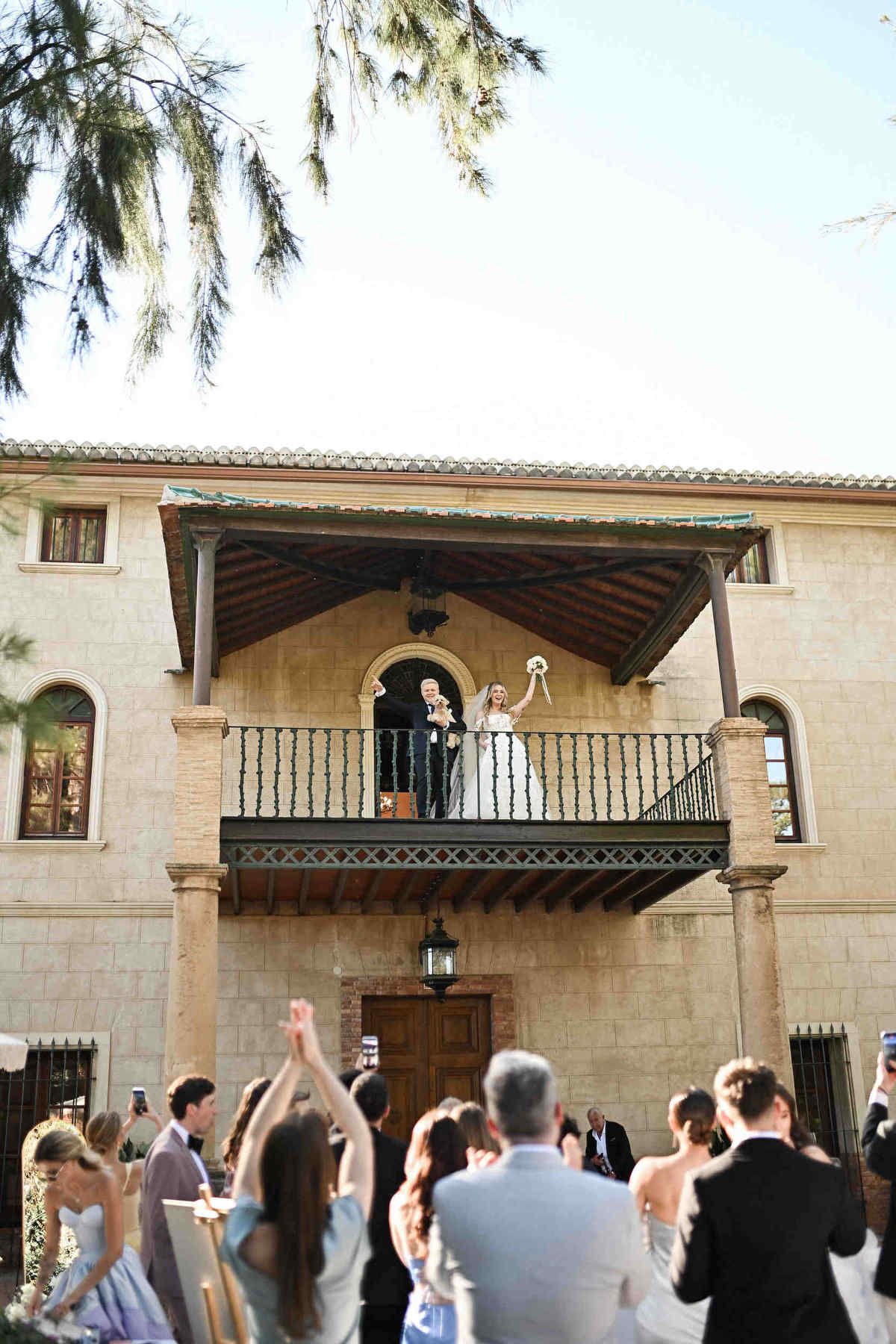 Newlyweds and their dog waving from the rustic balcony of Finca El Senyoret as wedding guests cheer below.