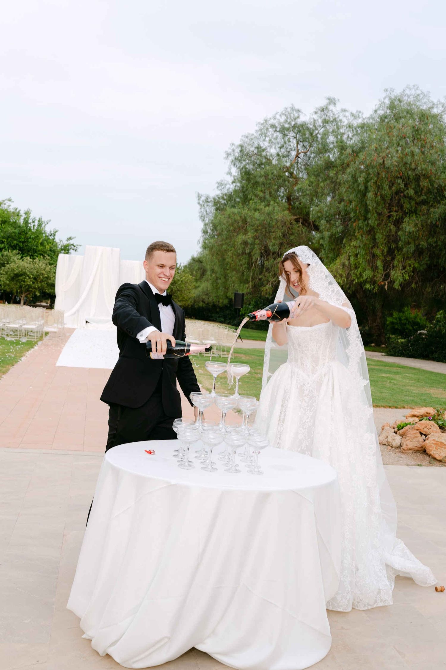 Groom in a tuxedo and bride in a lace veil pouring a champagne tower together on an outdoor stone terrace at a Spanish estate.