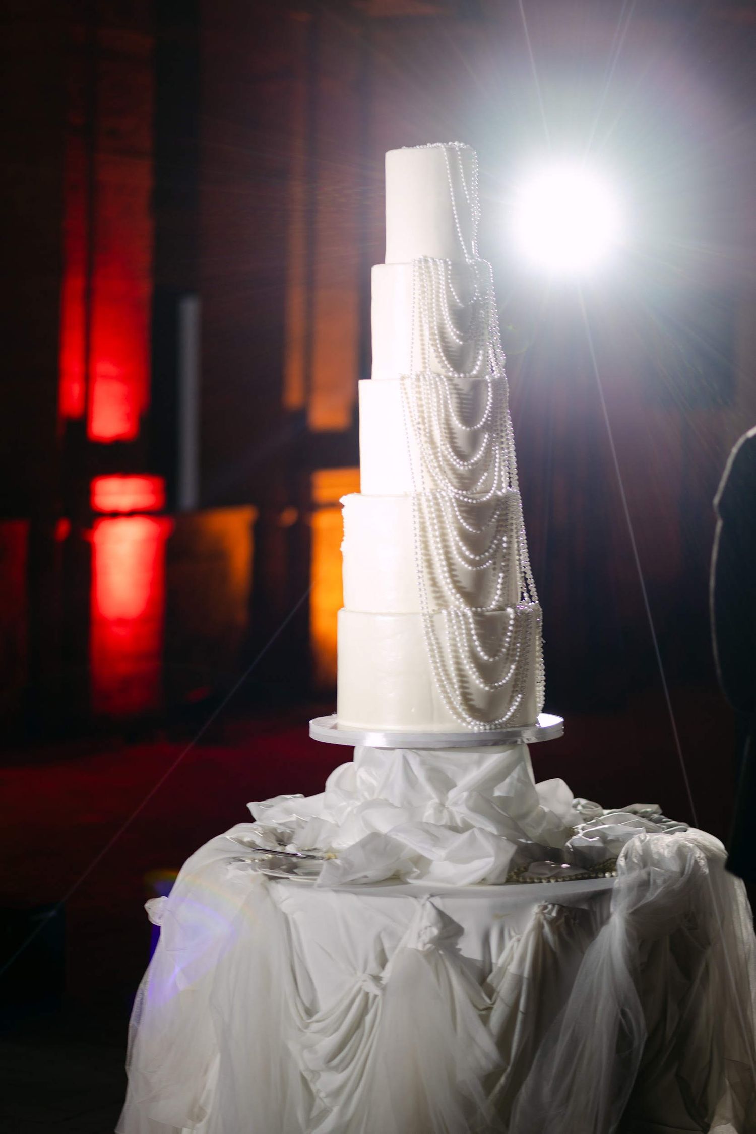 A tall multi-tiered white wedding cake draped in pearls stands at La Cartuja courtyard under red architectural lighting.
