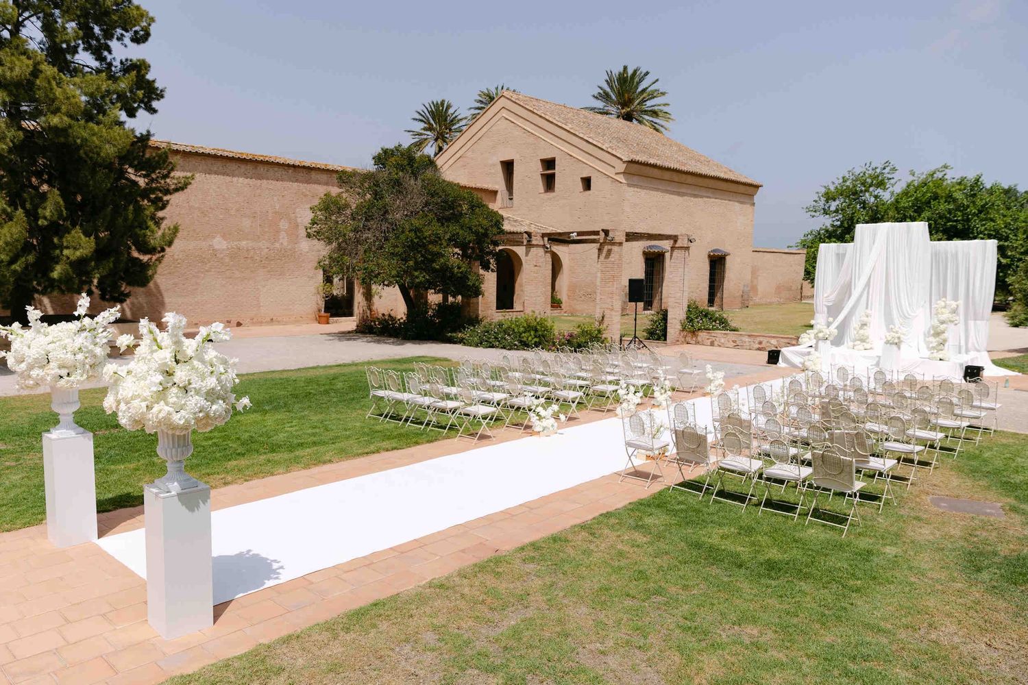 White vintage chairs lining a ceremony aisle on a green lawn outside the historic La Cartuja de Ara Christi monastery.