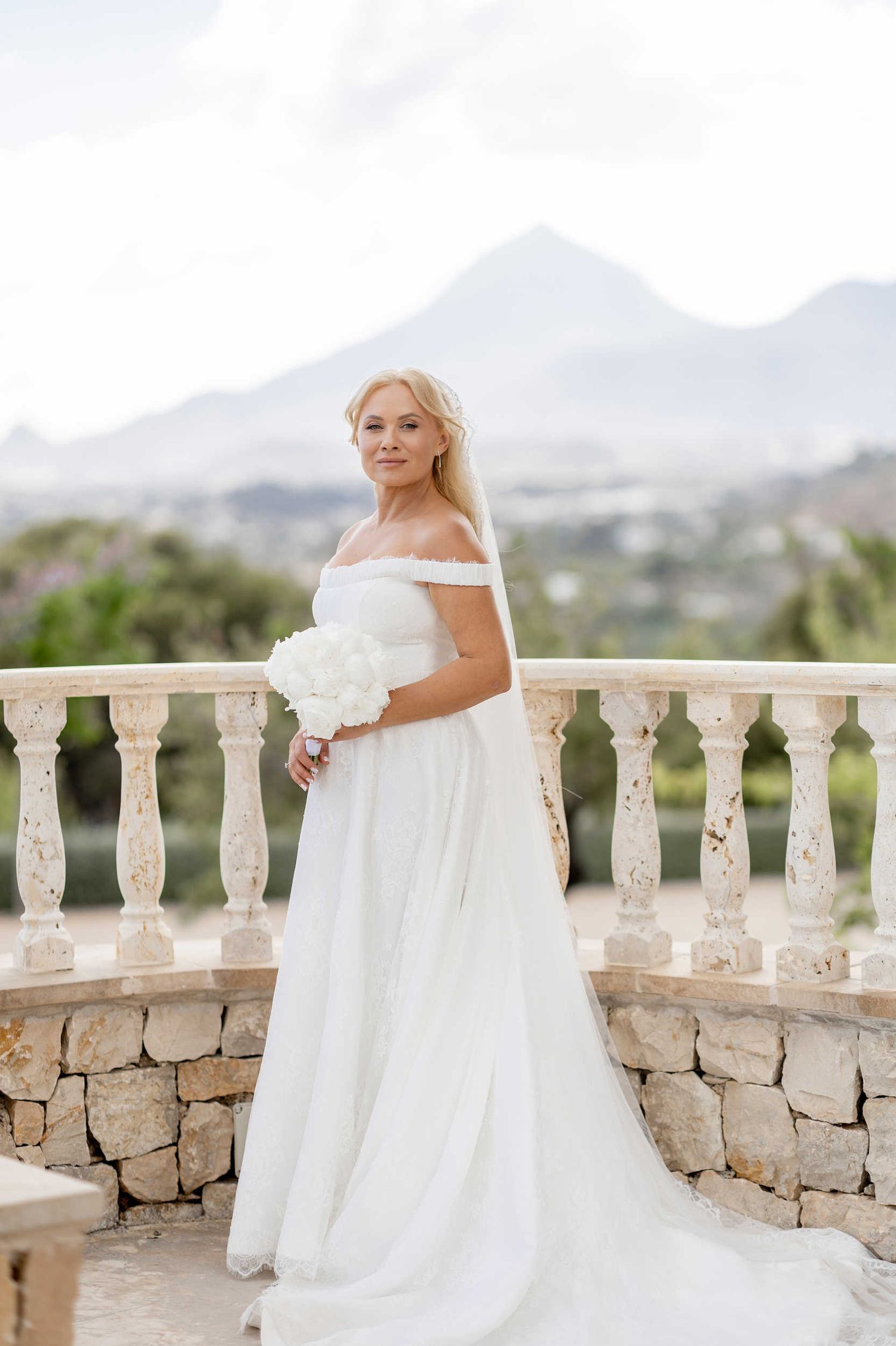 Beautiful bride holding a white bouquet, posing on a stone balcony overlooking mountains at Finca Marques de Montemolar in Altea.