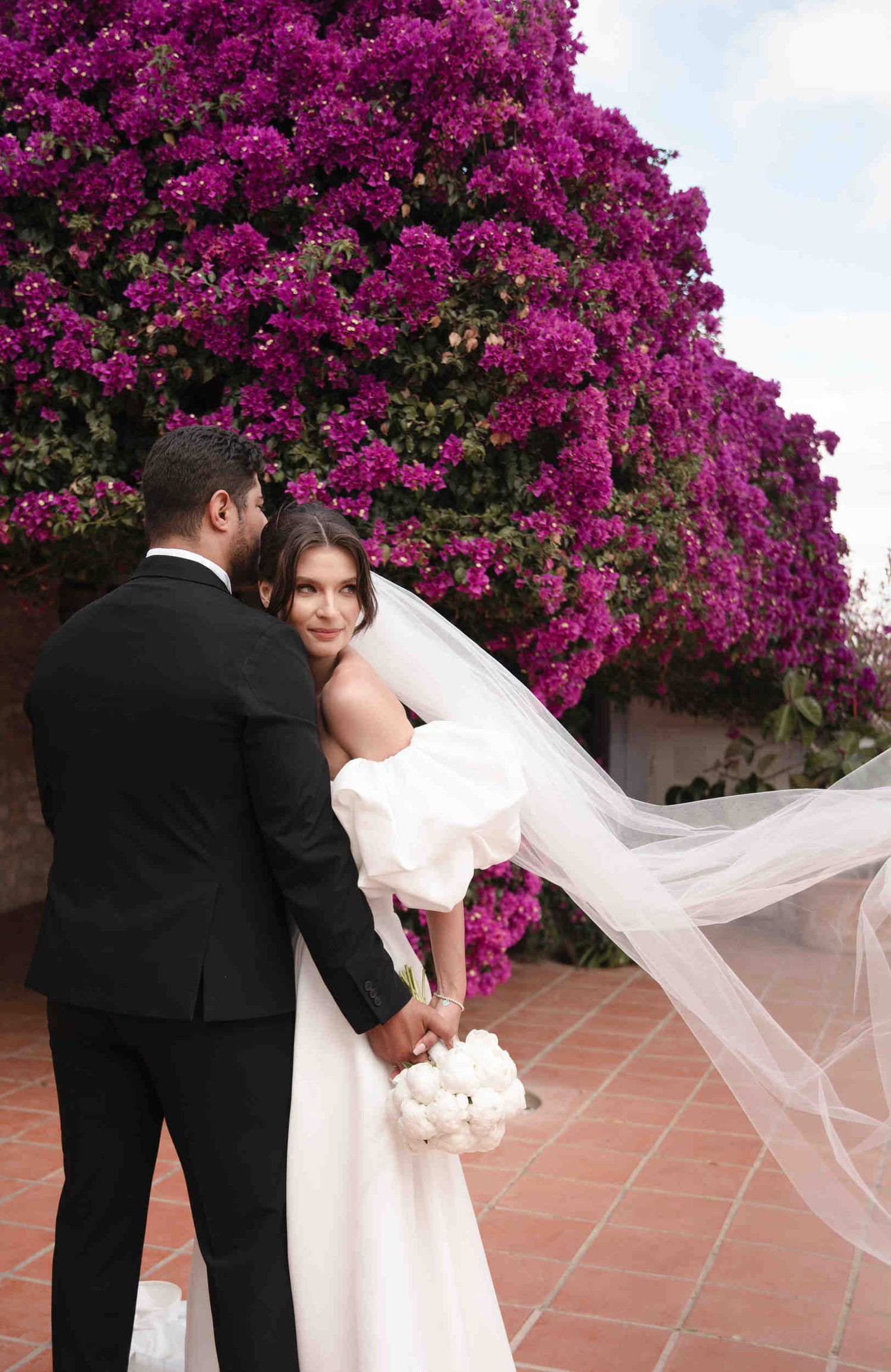 Destination wedding couple hugging in front of a vibrant purple bougainvillea bush at a sunny Valencia estate.