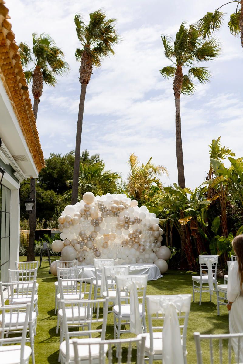 Empty white chairs face a large white and gold balloon arch under tall palm trees for an outdoor wedding ceremony in Alicante.