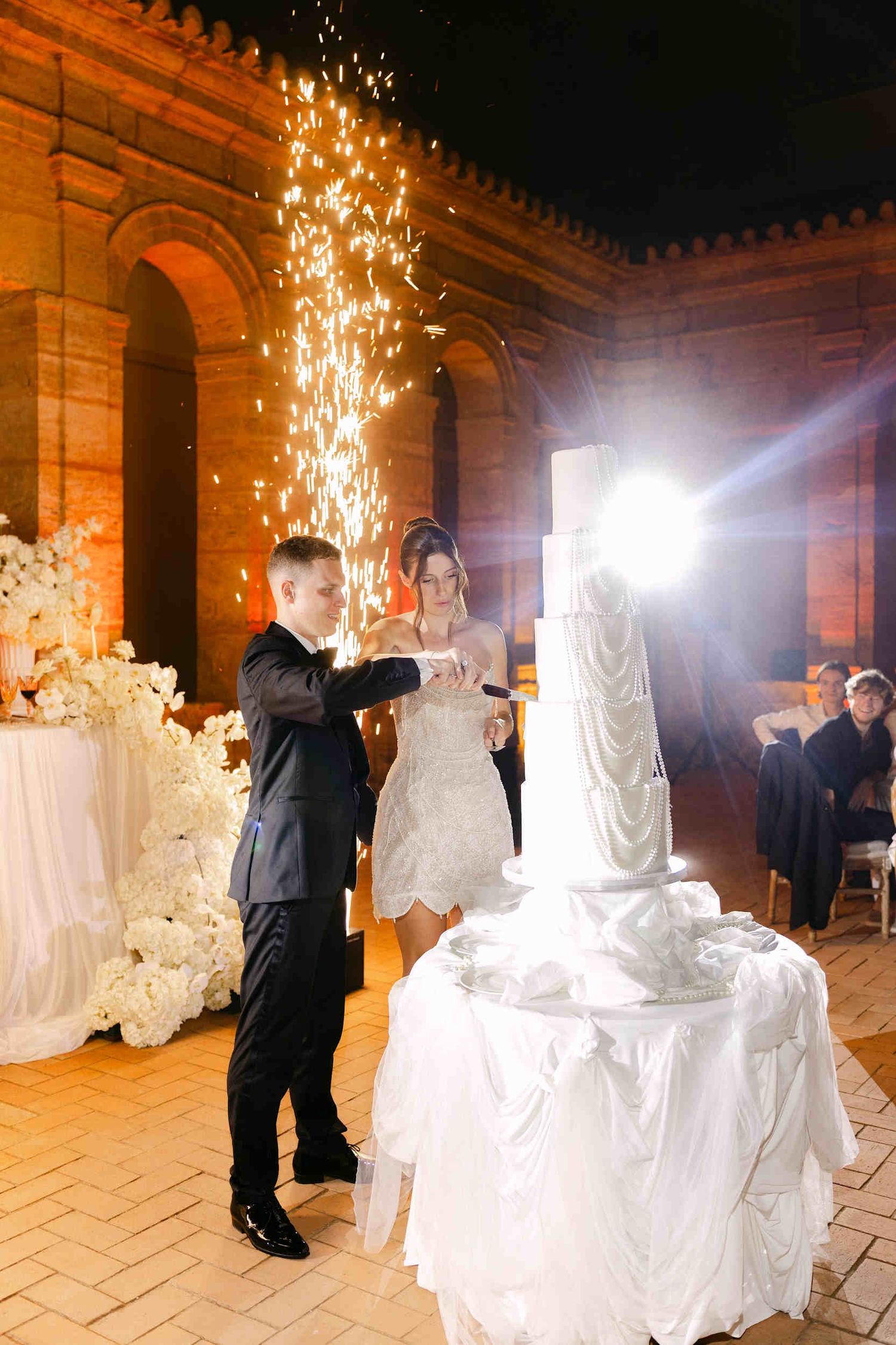 Bride and groom cutting a tall pearl wedding cake with bright sparklers in the stone courtyard of La Cartuja de Ara Christi.