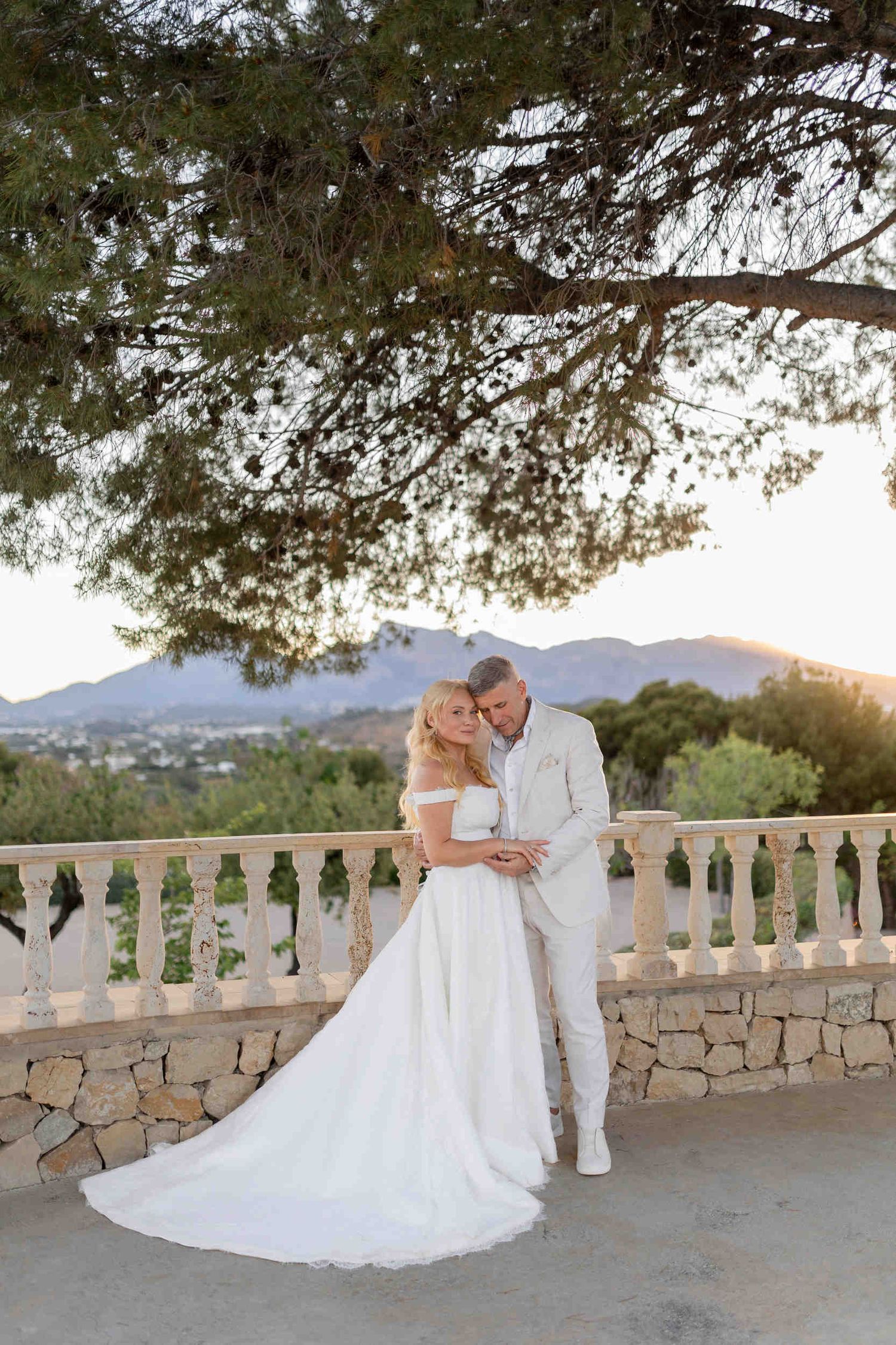 Mature wedding couple hugging on a stone terrace at sunset in Montemolar. Bride in white dress, groom in white suit.