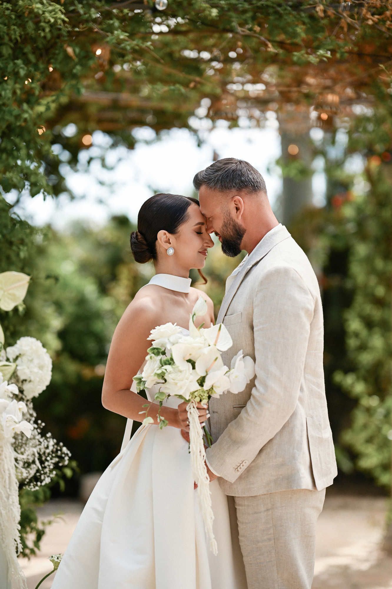 Romantic bride and groom touching foreheads under a floral arch with a white bouquet at their Villa Paula wedding.