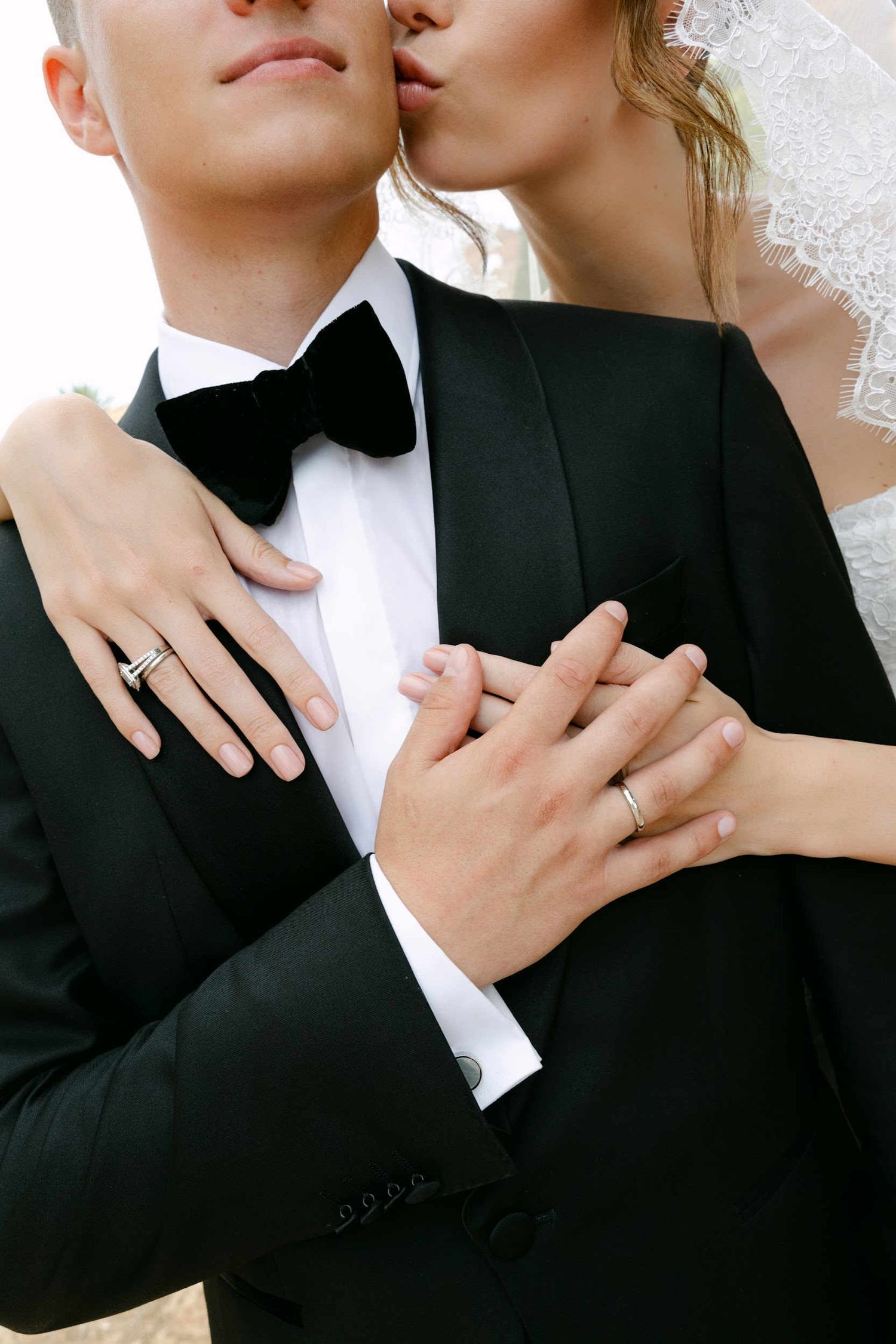 Close-up portrait of a bride kissing her groom on the cheek, highlighting her lace veil and wedding rings outdoors.