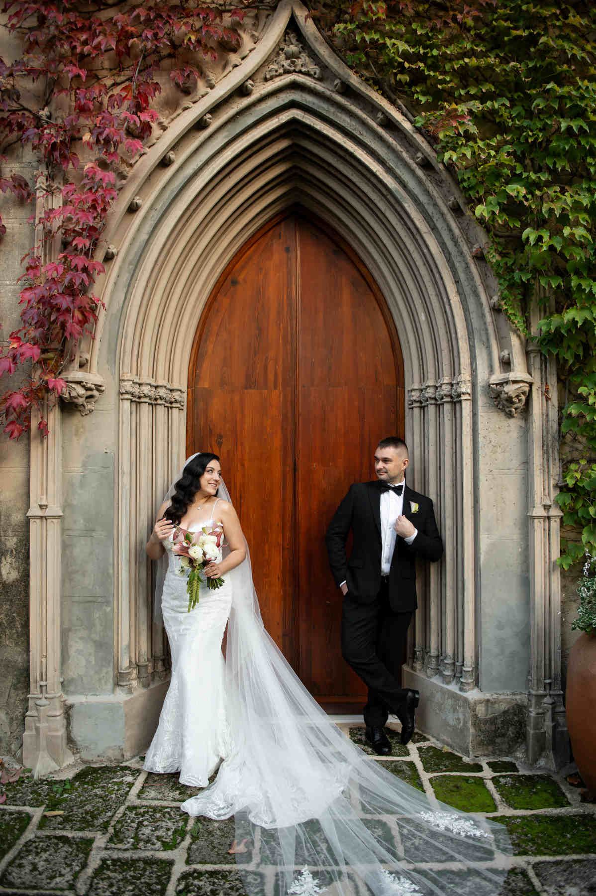 Newlyweds posing by grand wooden doors inside an ivy-framed stone archway at their beautiful Casa Santonja destination wedding.