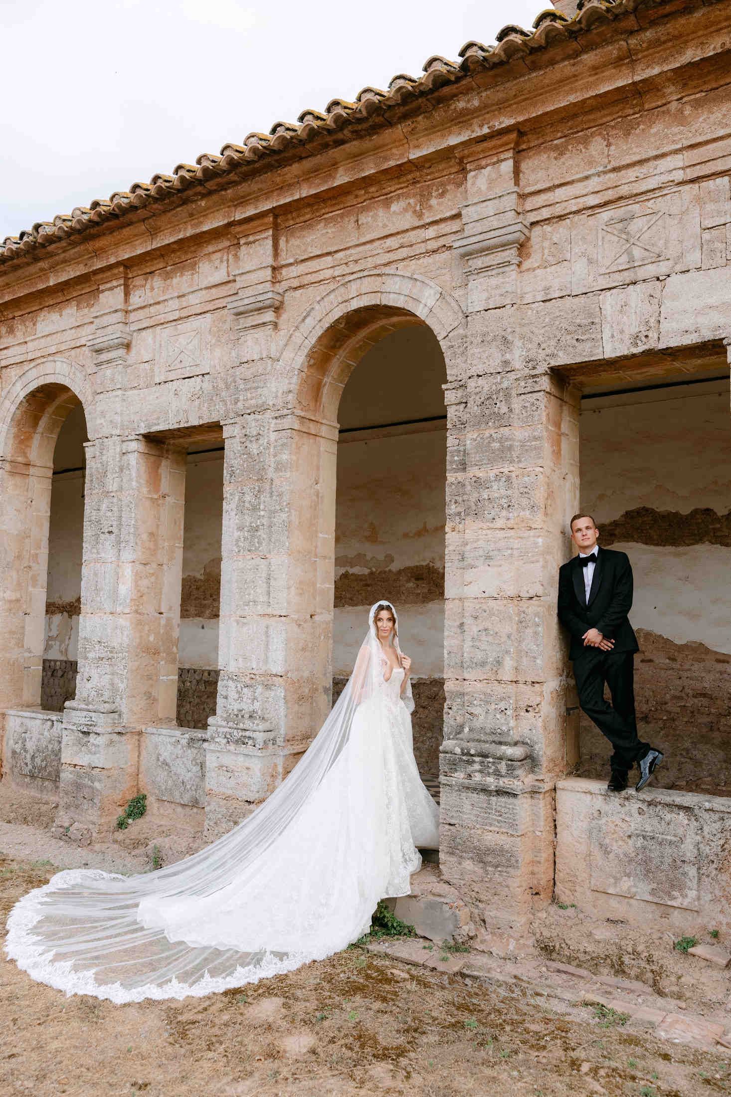 Bride wearing a long lace veil and groom in a black tuxedo posing apart in an ancient stone cloister ruin in Valencia.