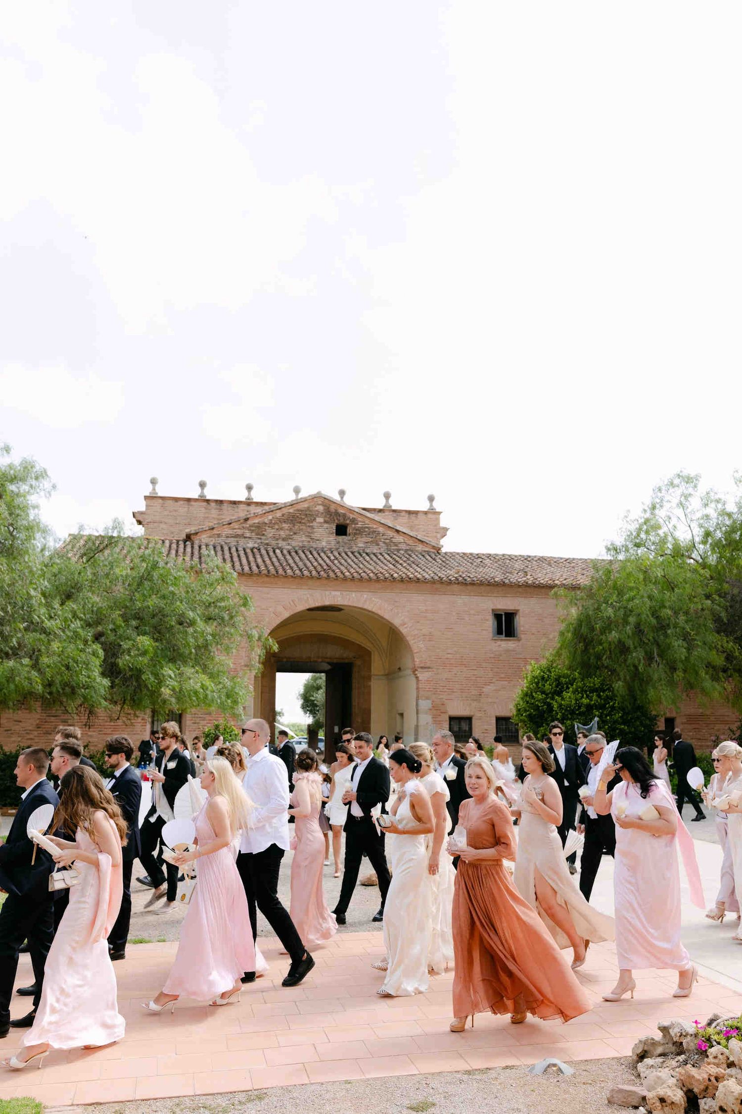 Wedding guests holding white paper fans walking across a sunlit stone courtyard at La Cartuja de Ara Christi.