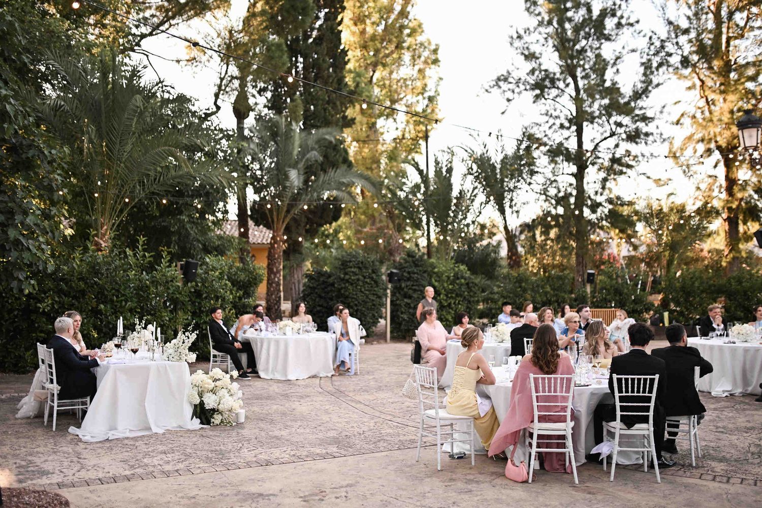 Guests and couple enjoying an outdoor wedding reception dinner at elegant round tables under pine trees and string lights in Benidorm