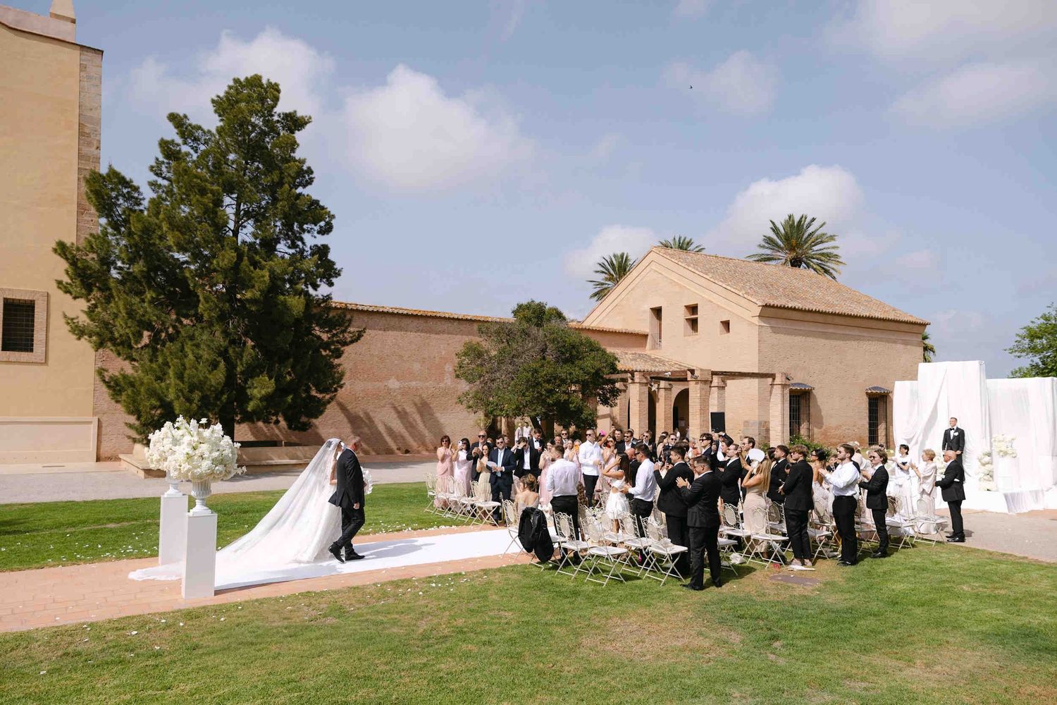 Guests stand and clap as the bride walks down a white aisle runner on a green lawn outside the historic Cartuja monastery.
