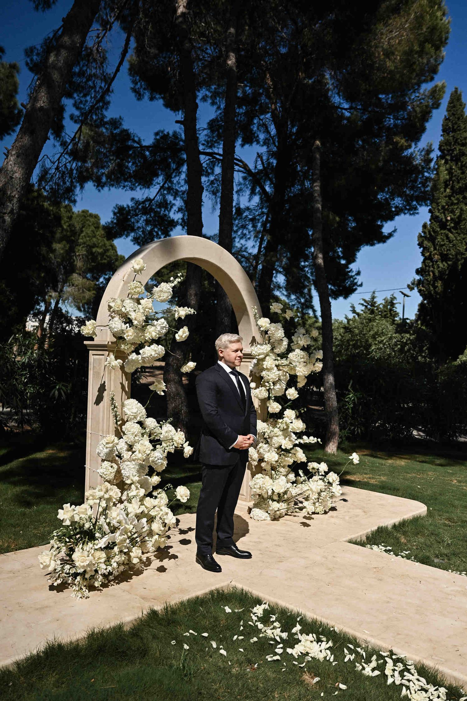 Groom waiting under a stone arch decorated with white roses before an outdoor wedding ceremony at Finca El Senyoret