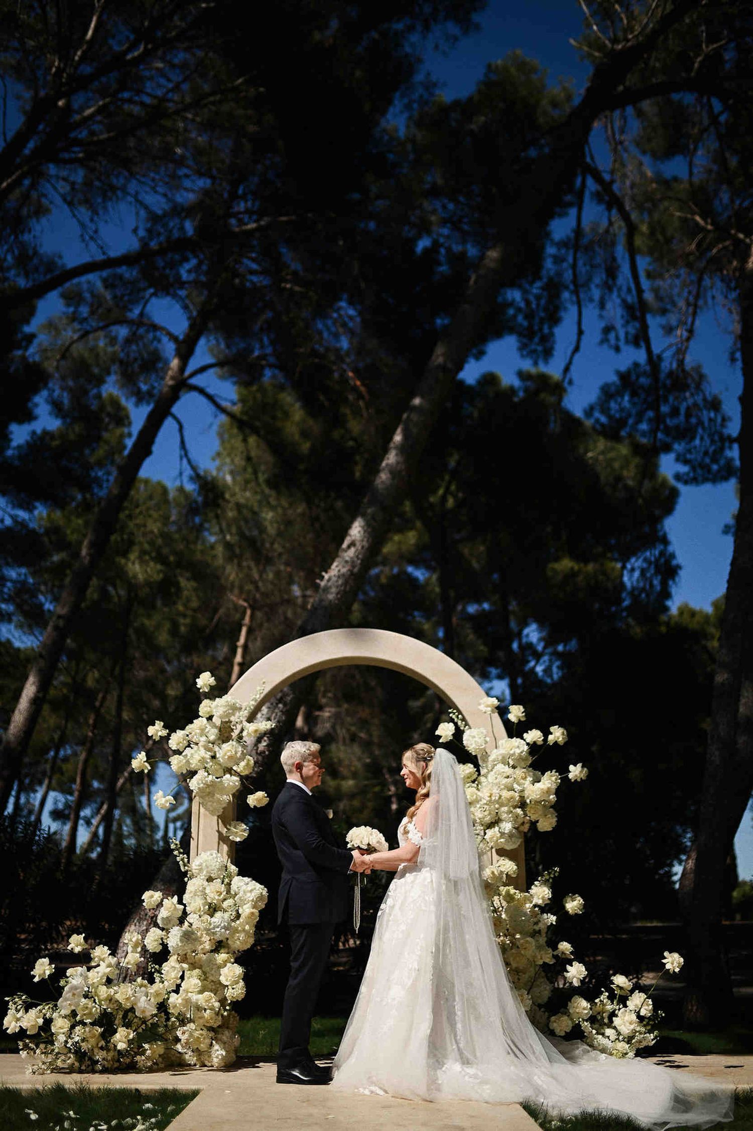 Bride and groom holding hands under a large floral stone arch during an outdoor pine forest wedding ceremony in Benidorm