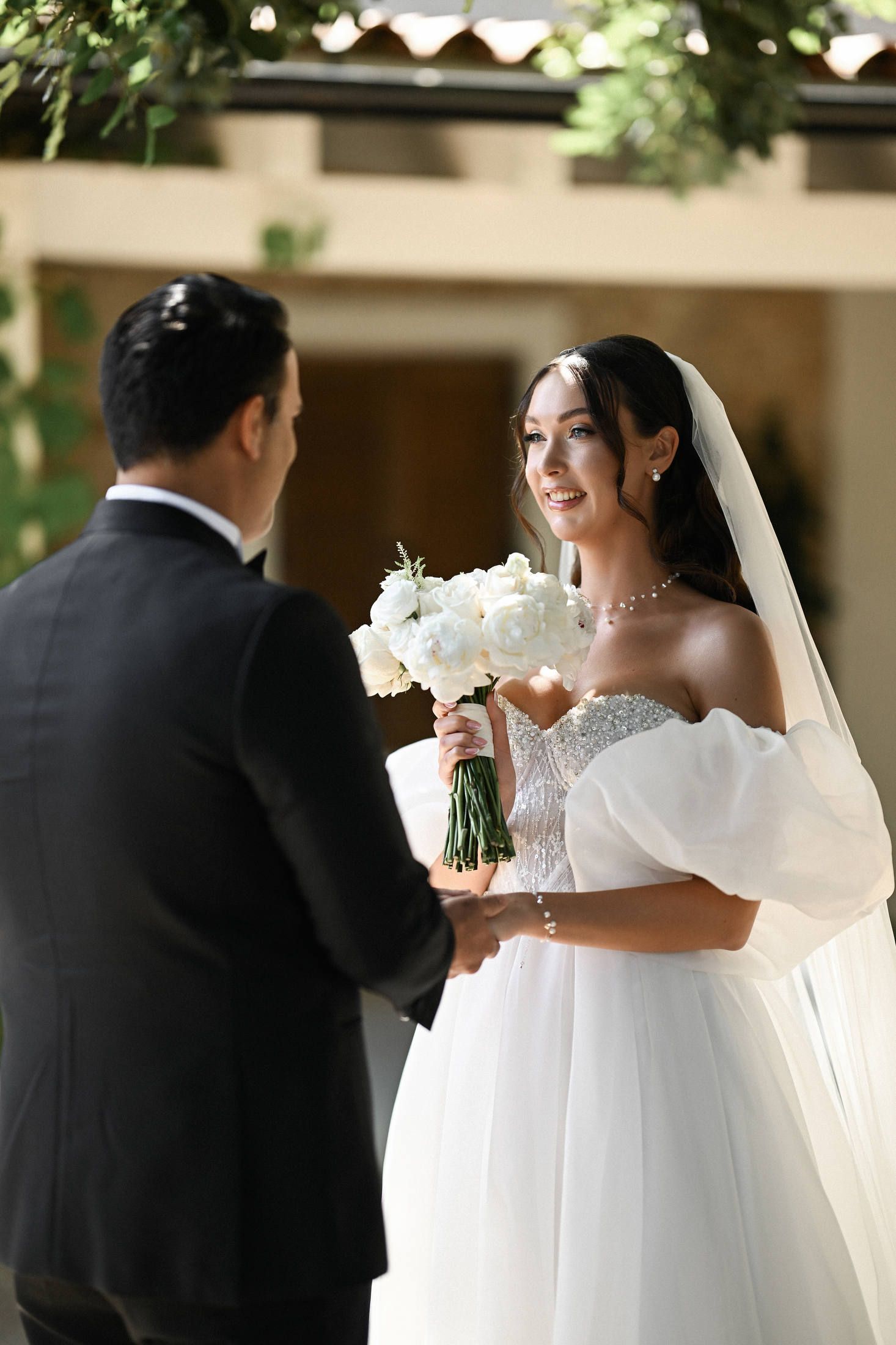 Bride in a puff-sleeve dress holding a white bouquet, smiling at her groom during their first look at a Valencia estate.