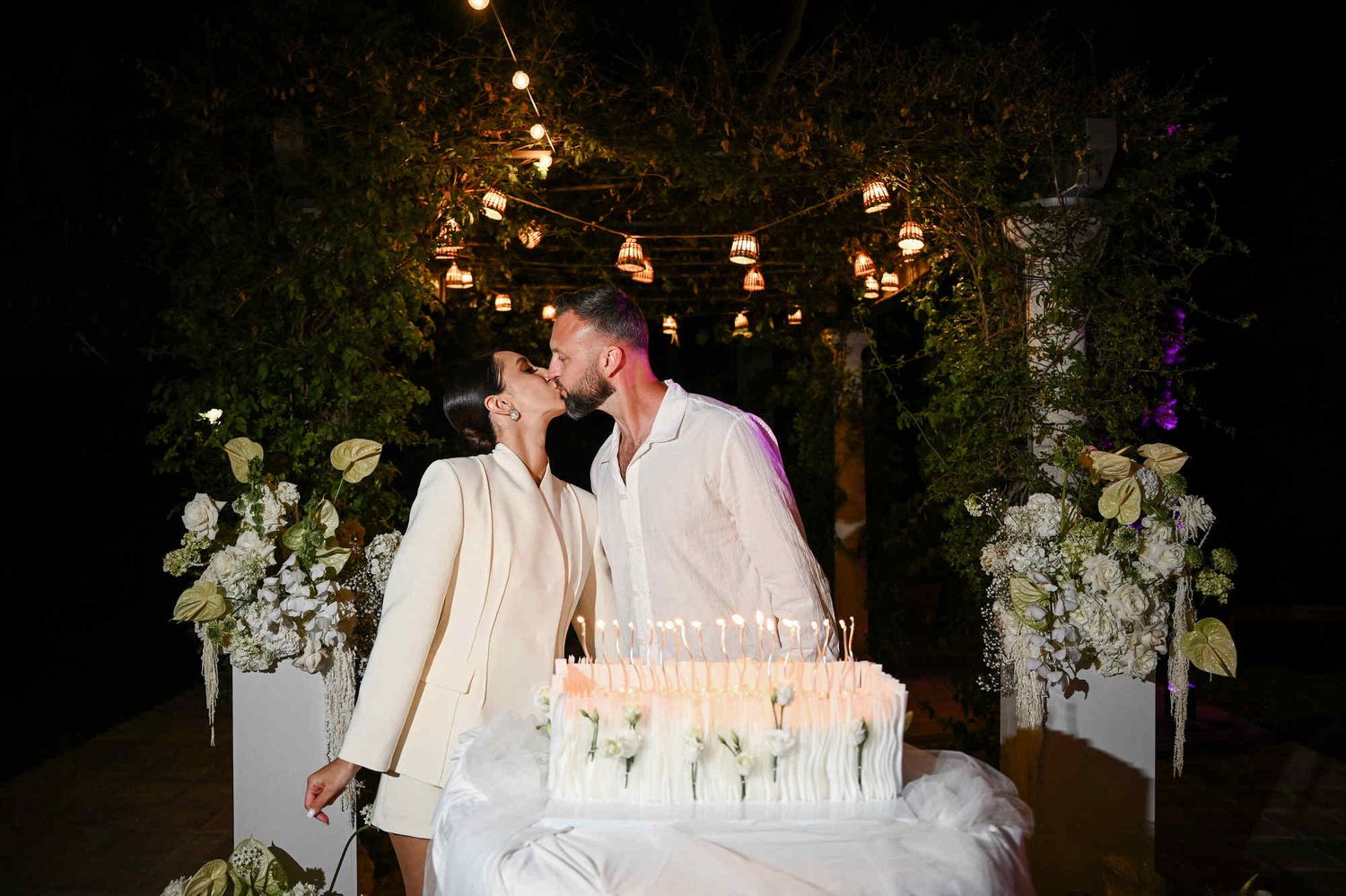 Bride and groom kissing behind an elegant white wedding cake lit with candles during their outdoor evening reception