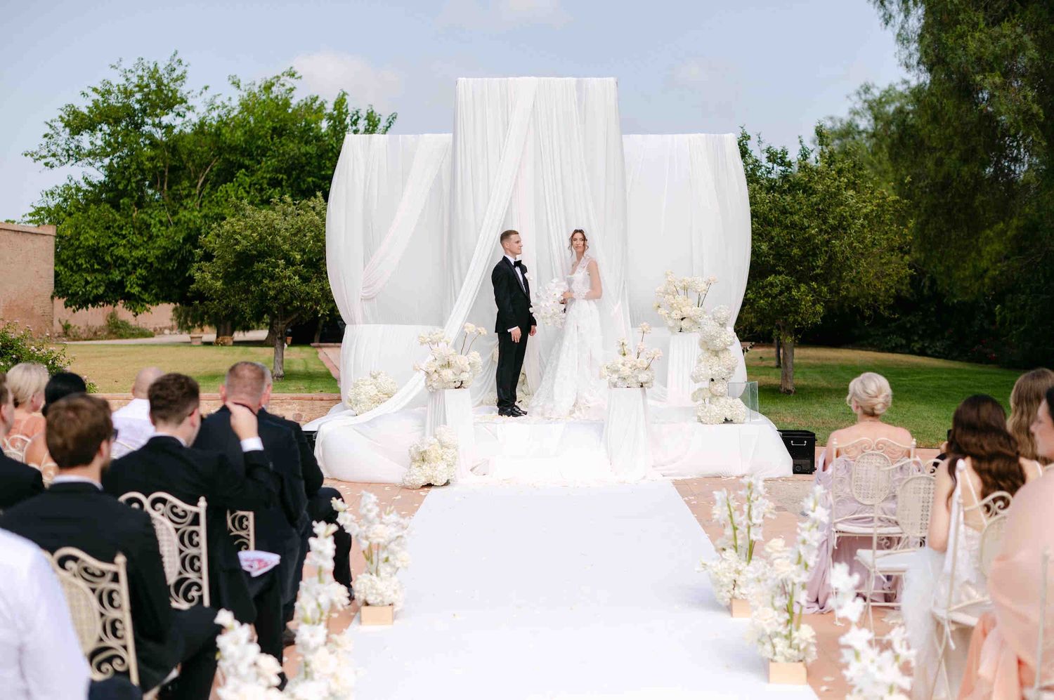Bride and groom stand together holding hands in front of a tall white fabric and floral altar at La Cartuja de Ara Christi.