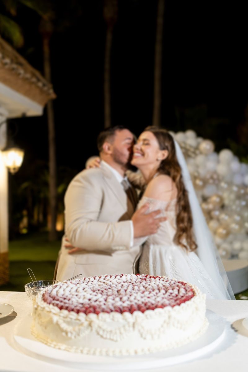 A raspberry wedding cake in sharp focus while a newlywed couple shares a kiss in the blurred background at an Alicante estate.