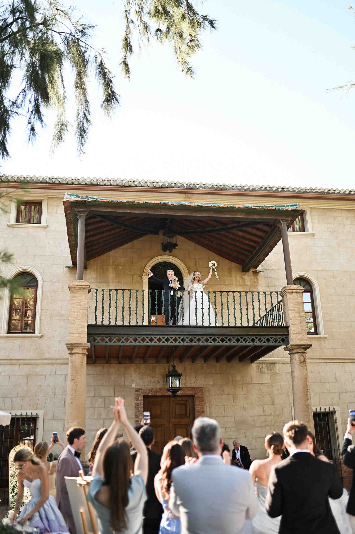Newlyweds holding a small dog and waving from a rustic balcony at a historic finca wedding in Benidorm.