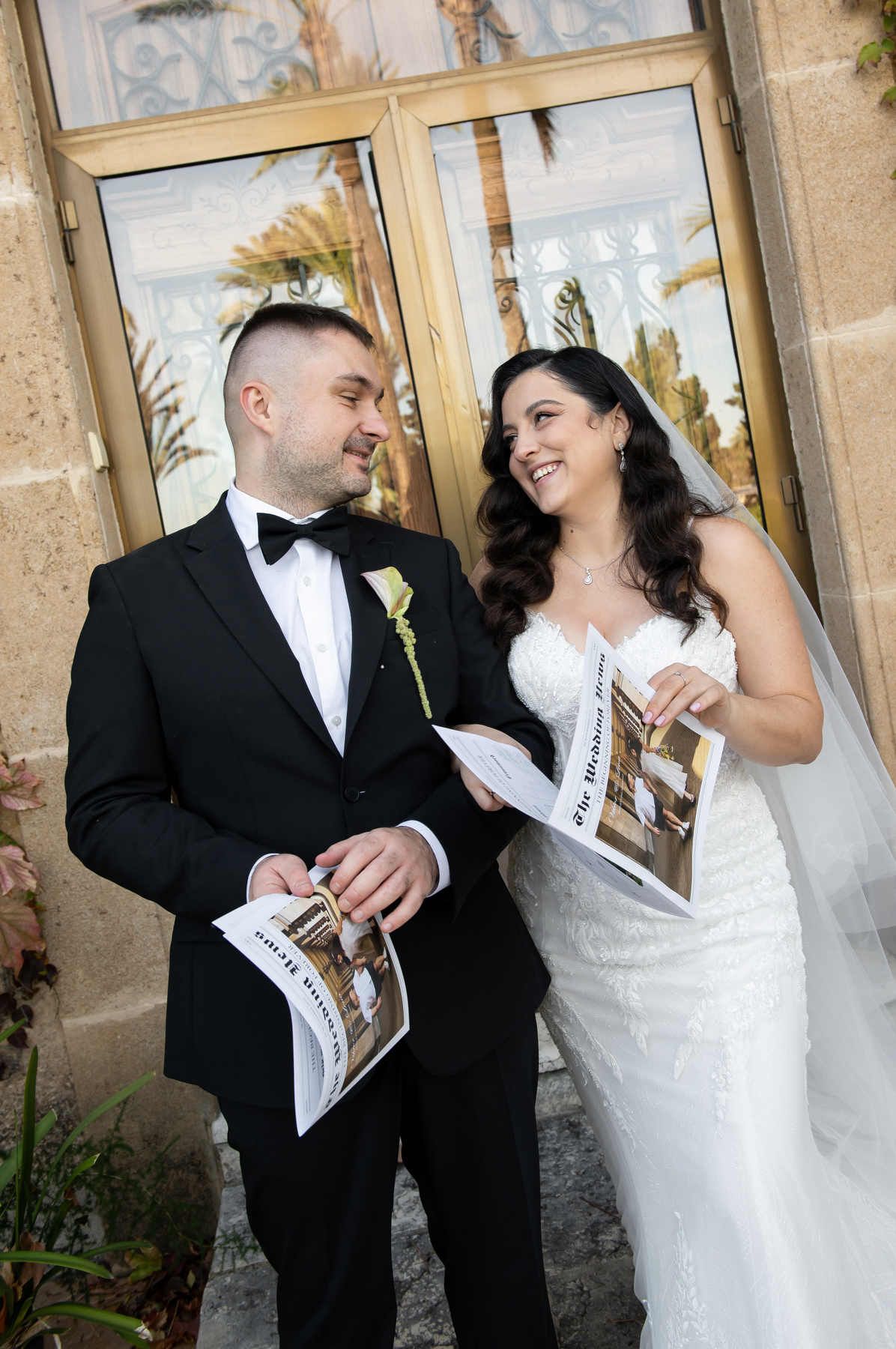Bride and groom laughing together while holding their personalized wedding newspaper outside the Casa Santonja estate.