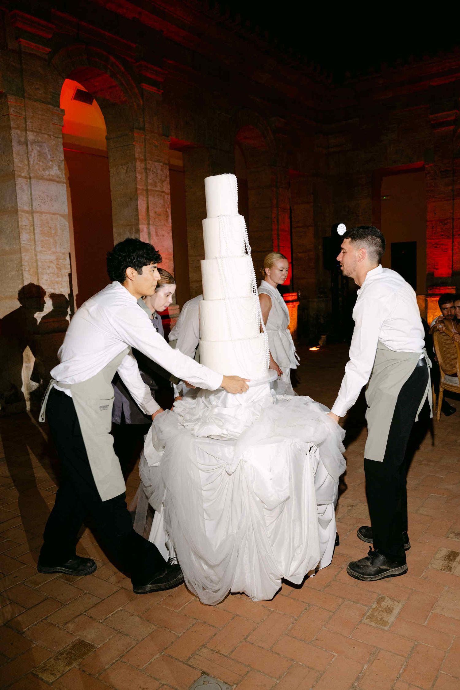Waiters carrying a massive multi-tiered white wedding cake decorated with pearls into the La Cartuja stone cloister courtyard.
