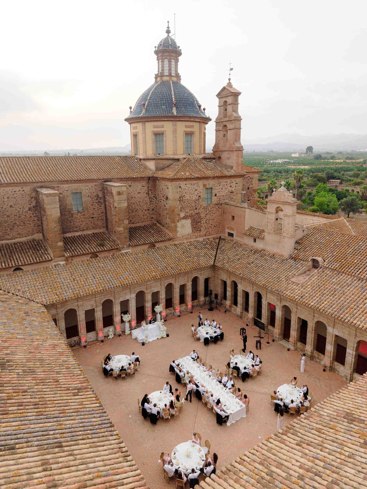 Wedding reception in la cartuja ara christi stone cloister featuring round tables, wooden chairs, and soft pink architectural uplighting.