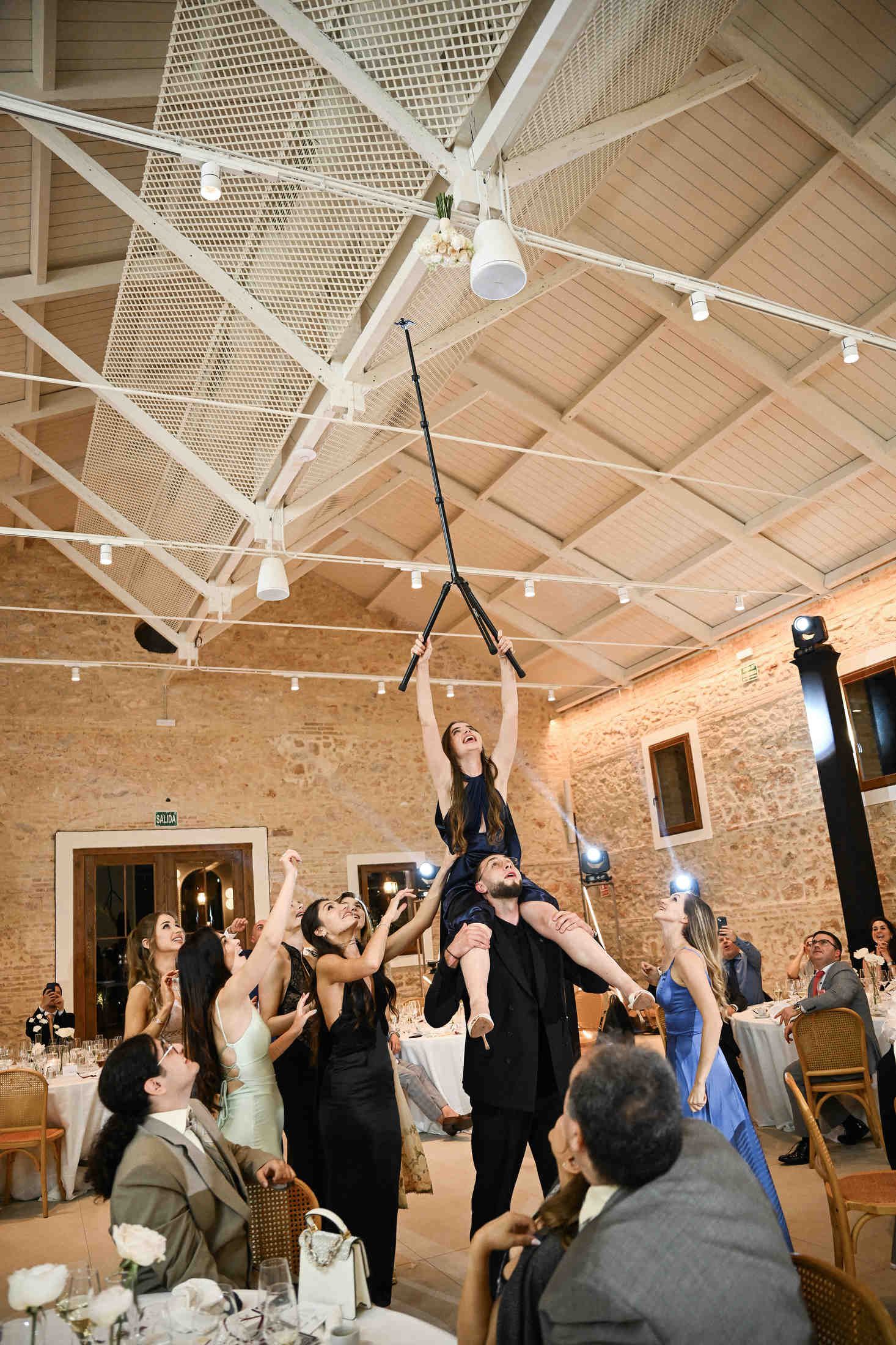 Wedding guests creatively using a tripod to rescue the bridal bouquet stuck on the ceiling at a finca el canonigo reception.