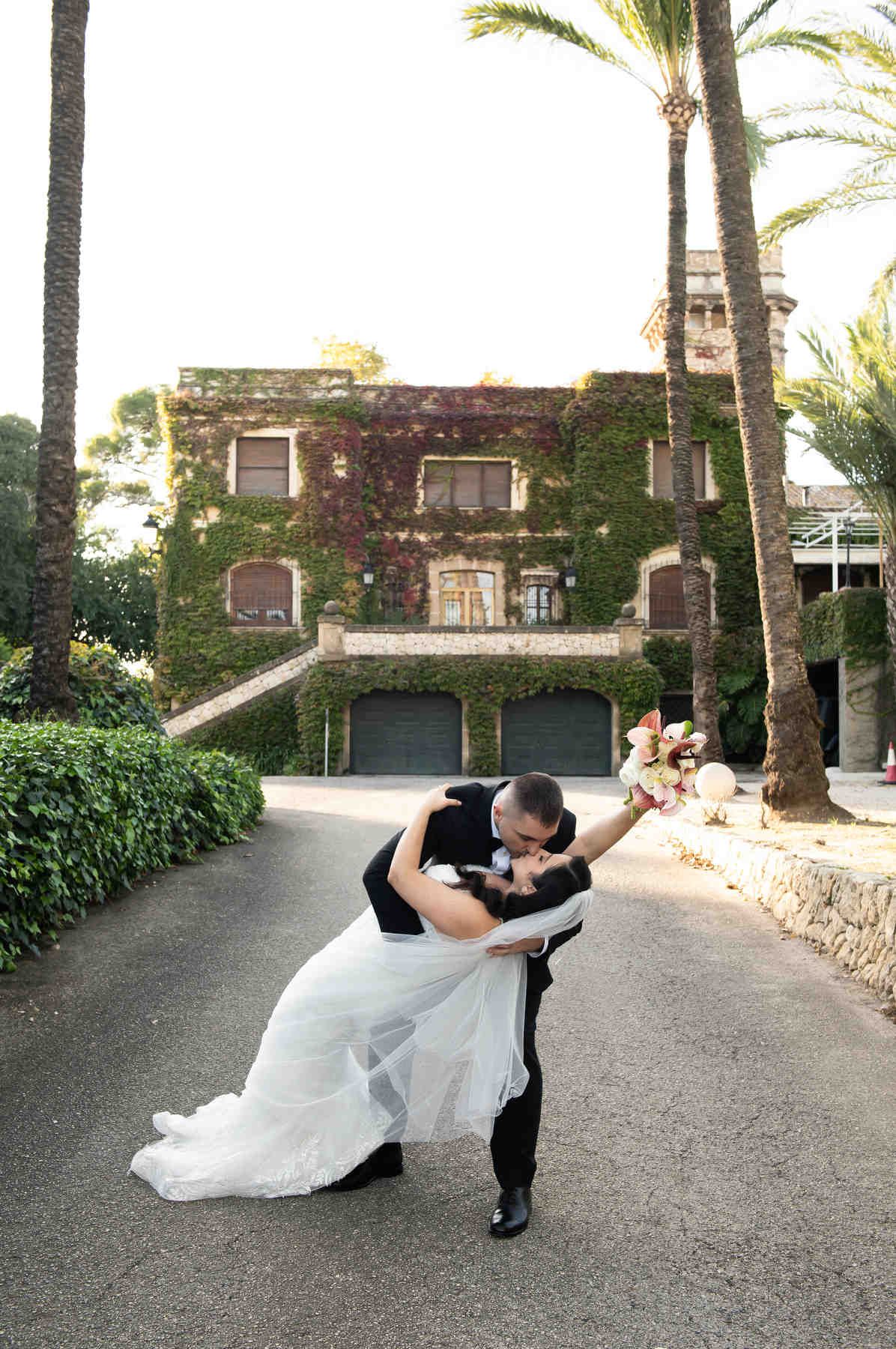 Groom dipping and kissing his bride on the driveway of the 19th-century Casa Santonja estate with red autumn vines and palm trees.