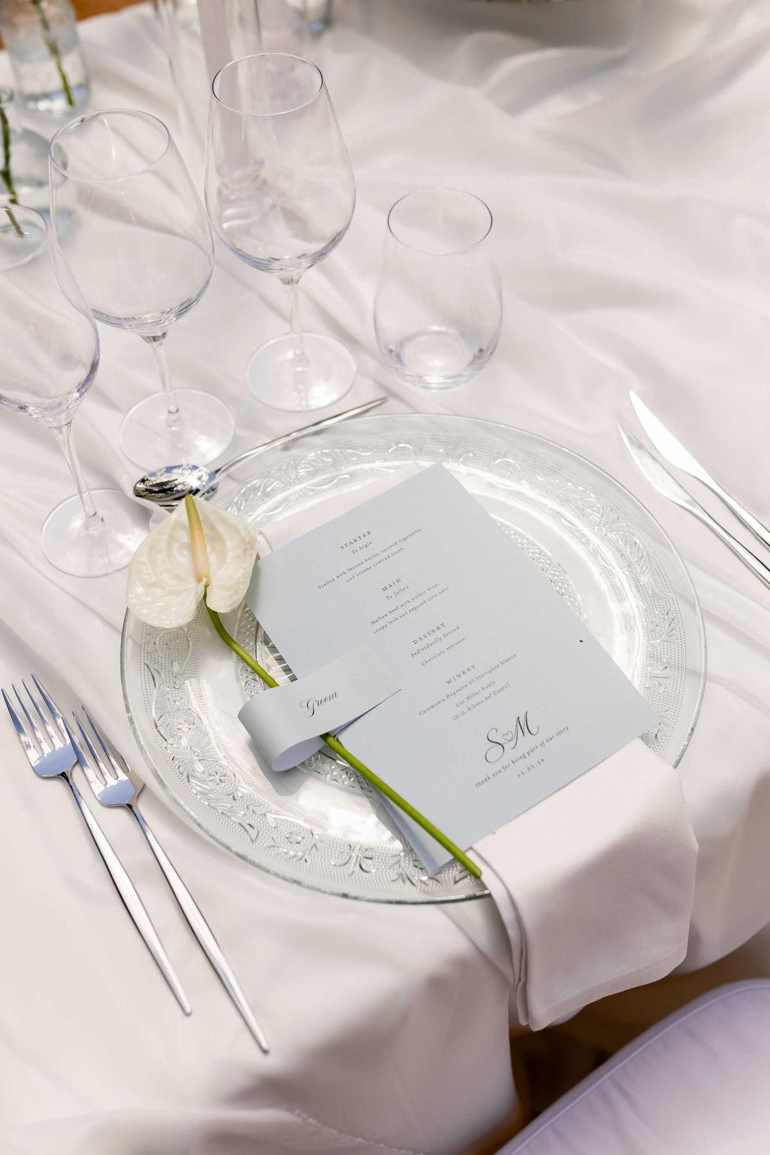Elegant wedding reception table setting with a light blue menu, crystal glassware, and white anthurium flower in Altea, Spain.