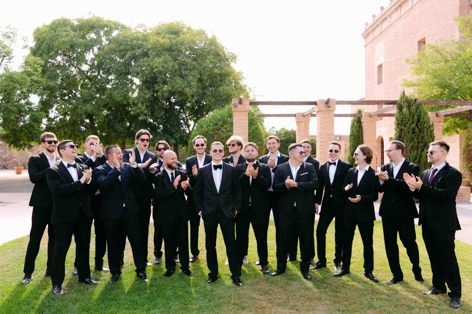 Groomsmen in black suits and sunglasses clapping on a green lawn outside a historic brick countryside estate in Valencia.