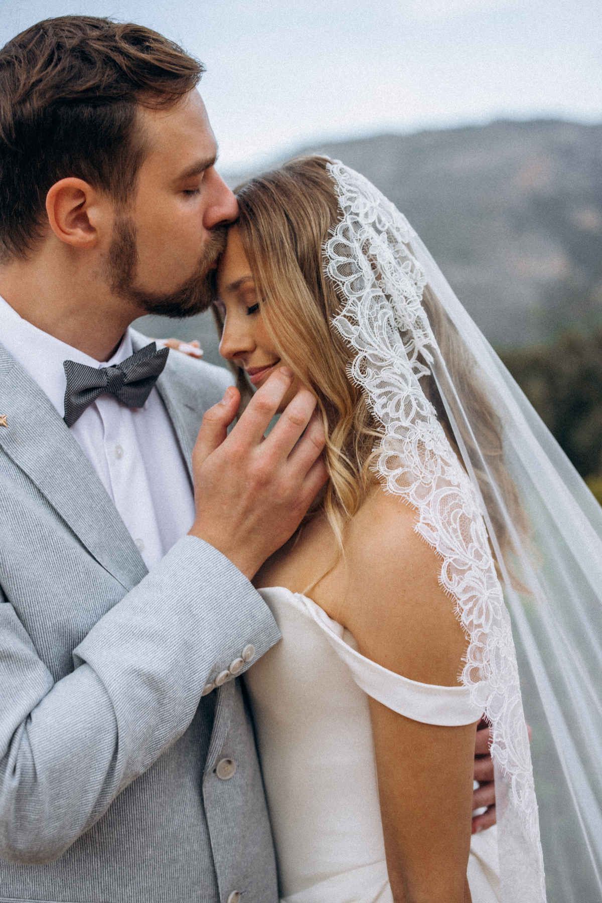 Close-up of a groom tenderly kissing his bride's forehead, highlighting her elegant lace veil at their destination wedding.