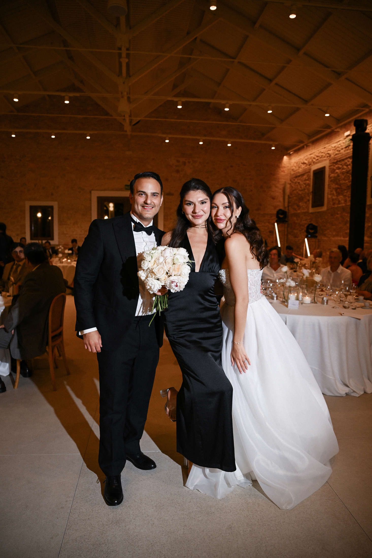 Bride and groom posing with a smiling guest in a black dress during their elegant indoor wedding reception in finca del canonigo