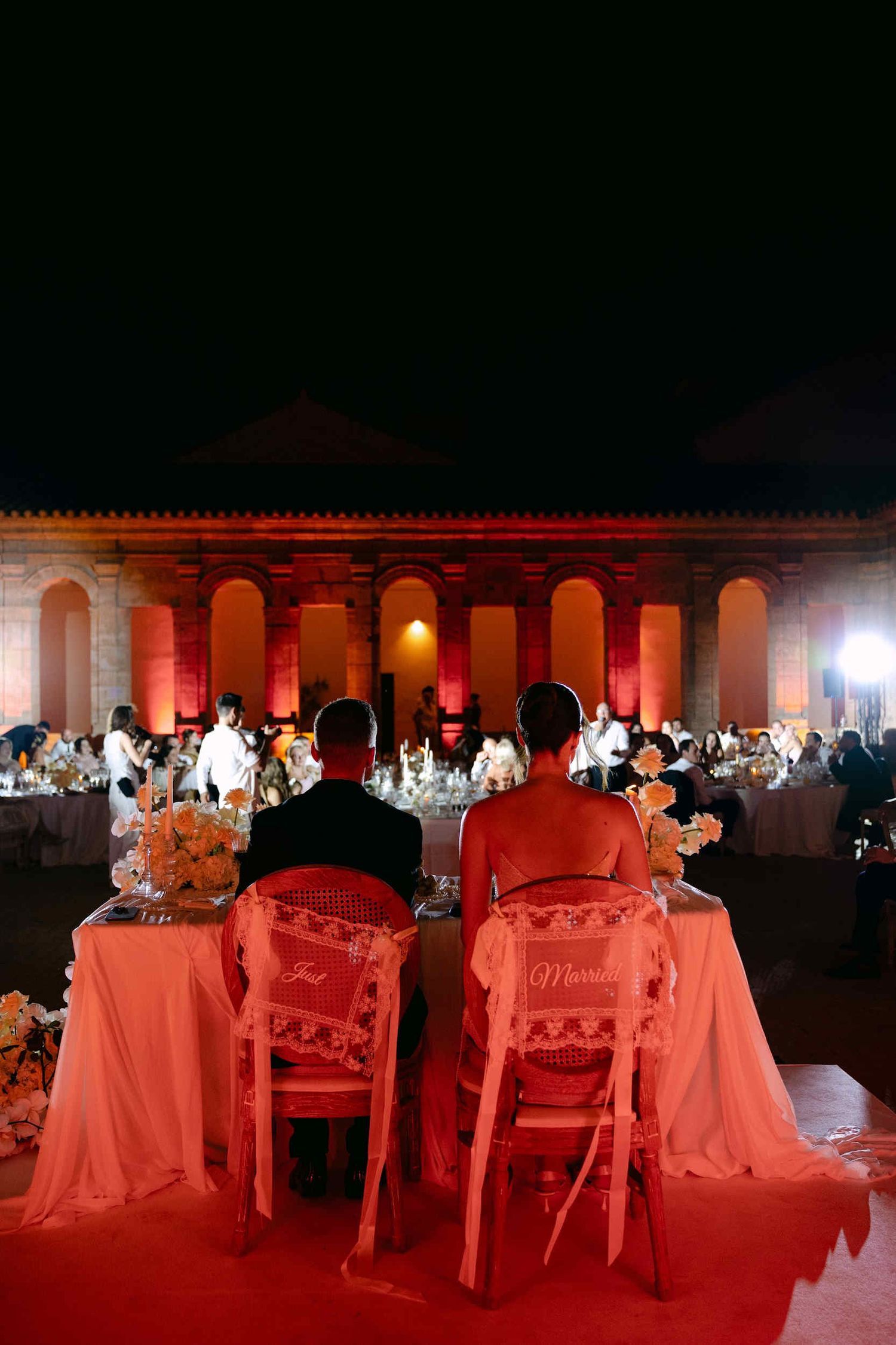 Newlyweds at a sweetheart table with Just Married signs during a reception at La Cartuja de Ara Christi under red lighting.
