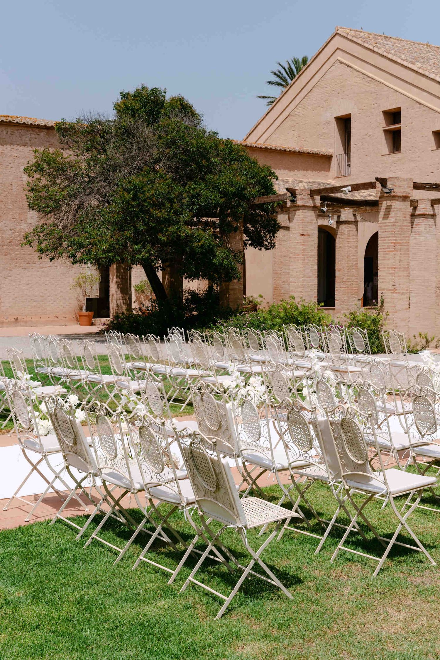 White vintage iron chairs lining a ceremony aisle outside a historic stone monastery building under bright sunlight.