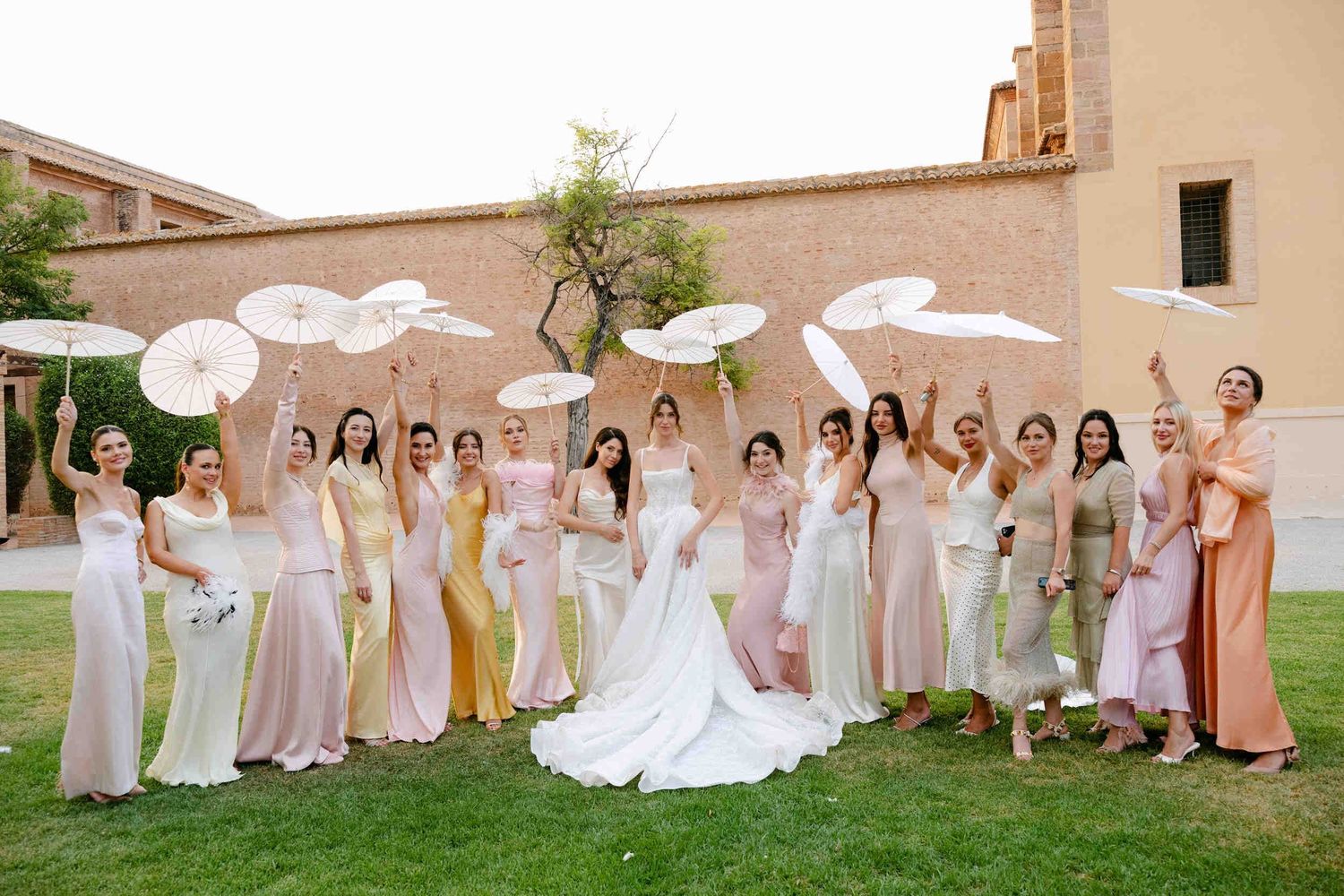 Bride surrounded by bridesmaids in pastel dresses holding white paper parasols on a green lawn at La Cartuja.