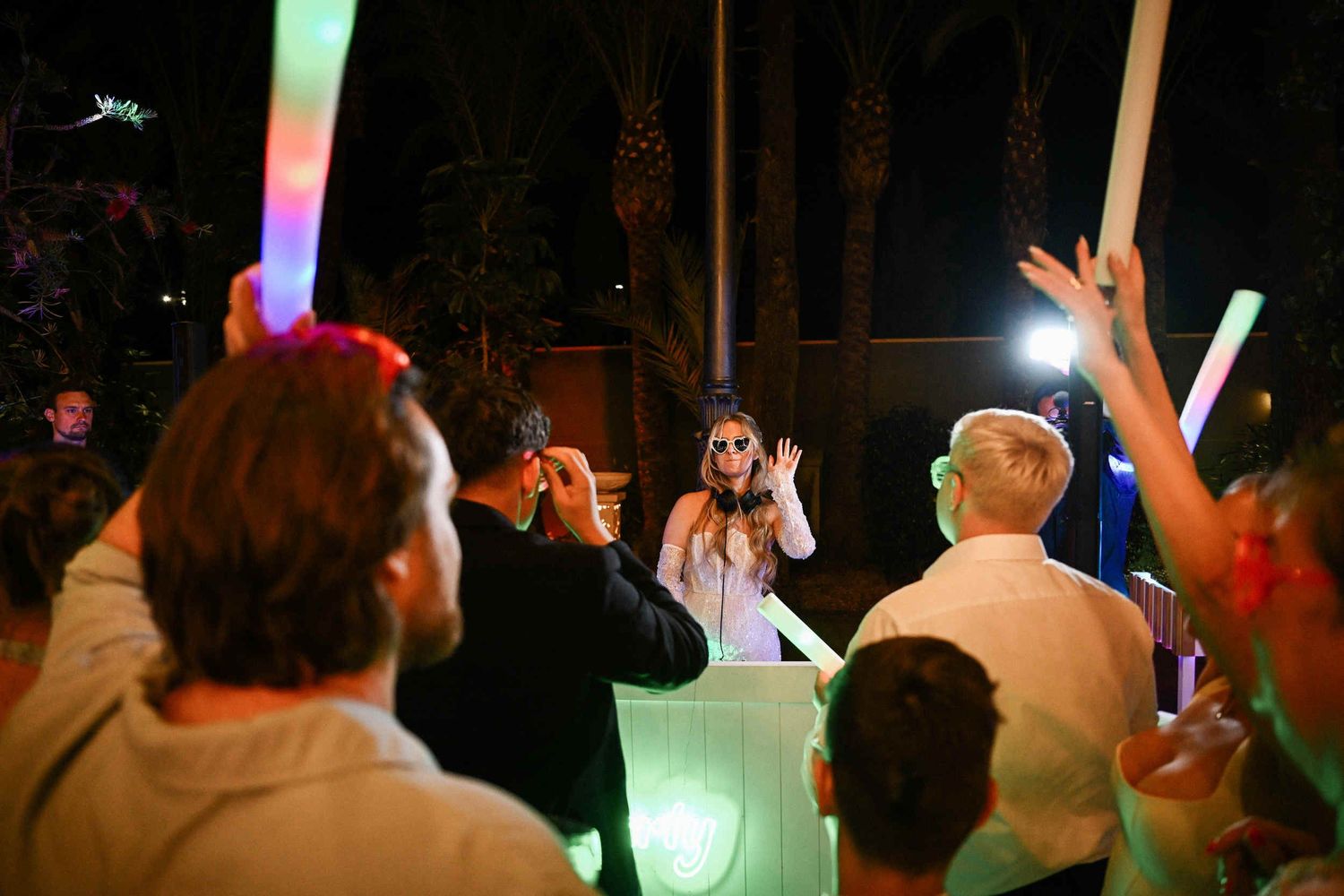 Bride wearing fun sunglasses and headphones DJing her own wedding party while guests cheer with glow sticks at a Spanish finca.