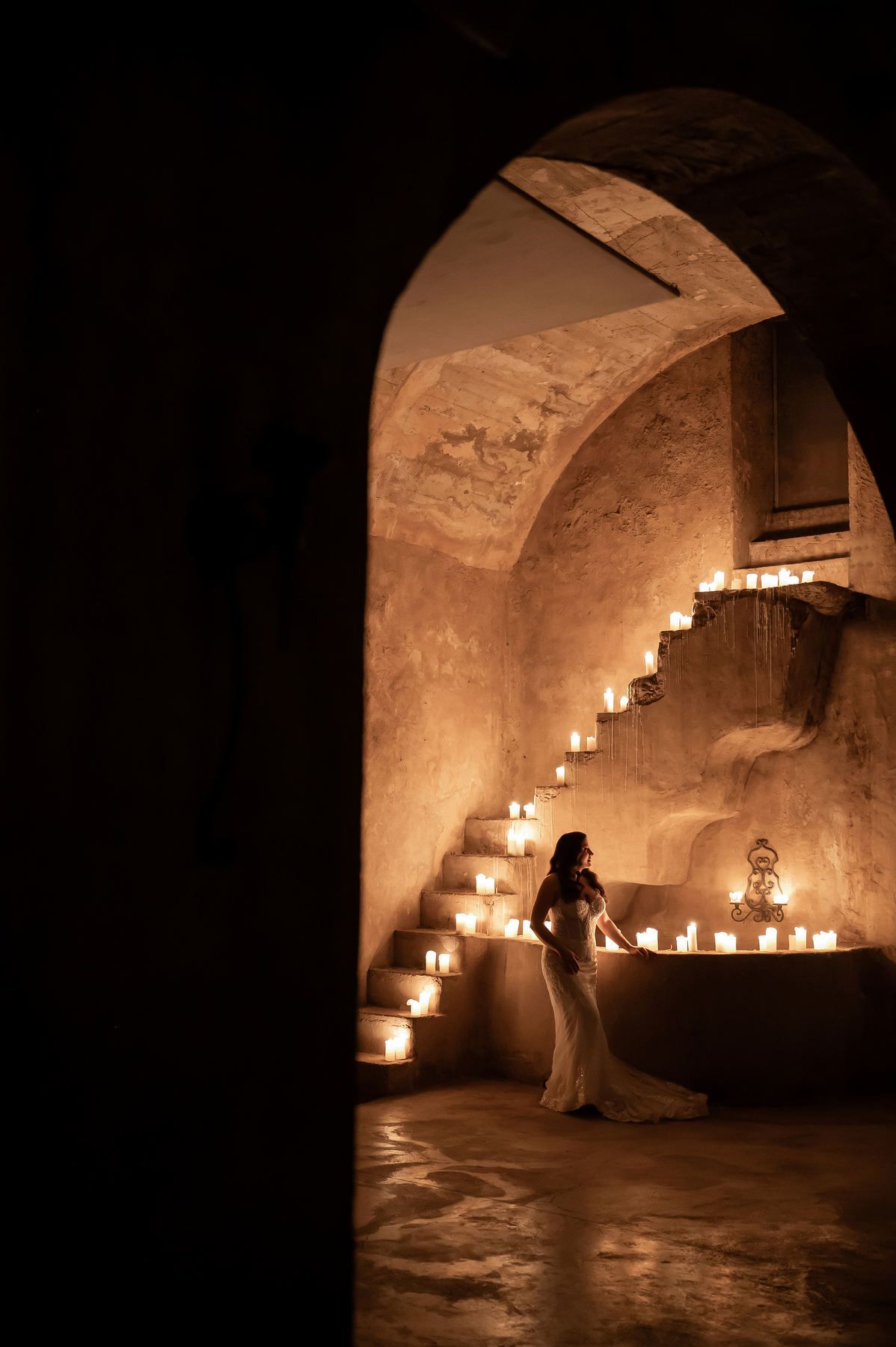 Framed view through a stone archway of a bride standing in a vaulted room illuminated by candles at Casa Santonja.