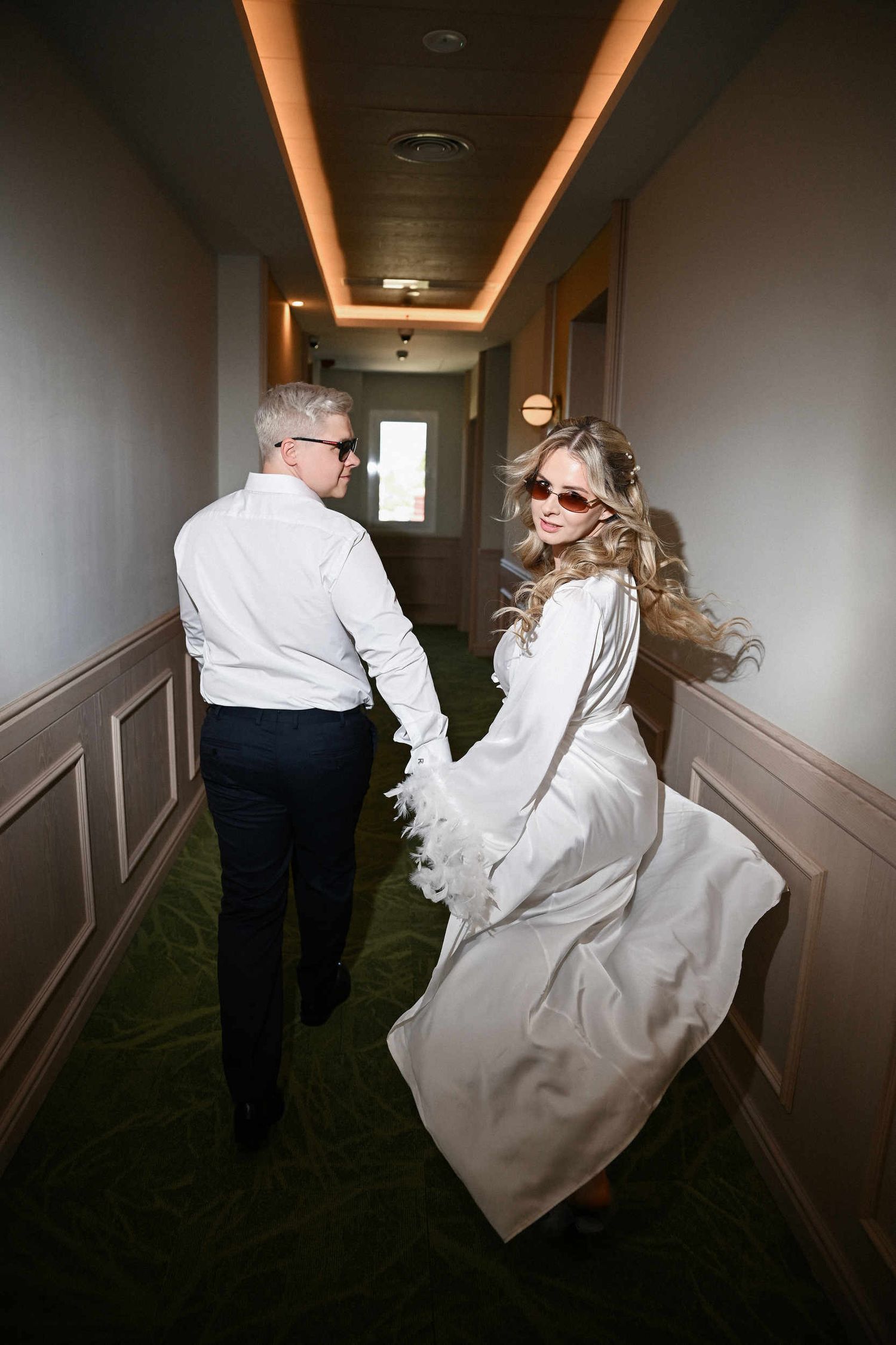 Bride and groom wearing sunglasses walking down a hallway, bride in a white feather robe before their benidorm wedding.