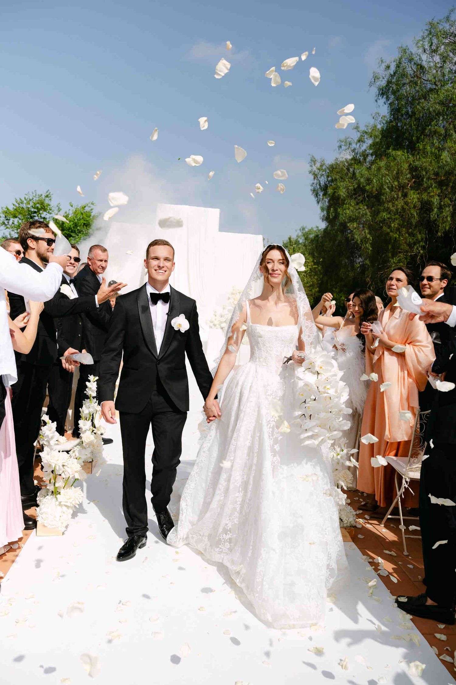 Bride and groom walking down a white aisle under rose petals. Joyful outdoor wedding ceremony in Valencia.