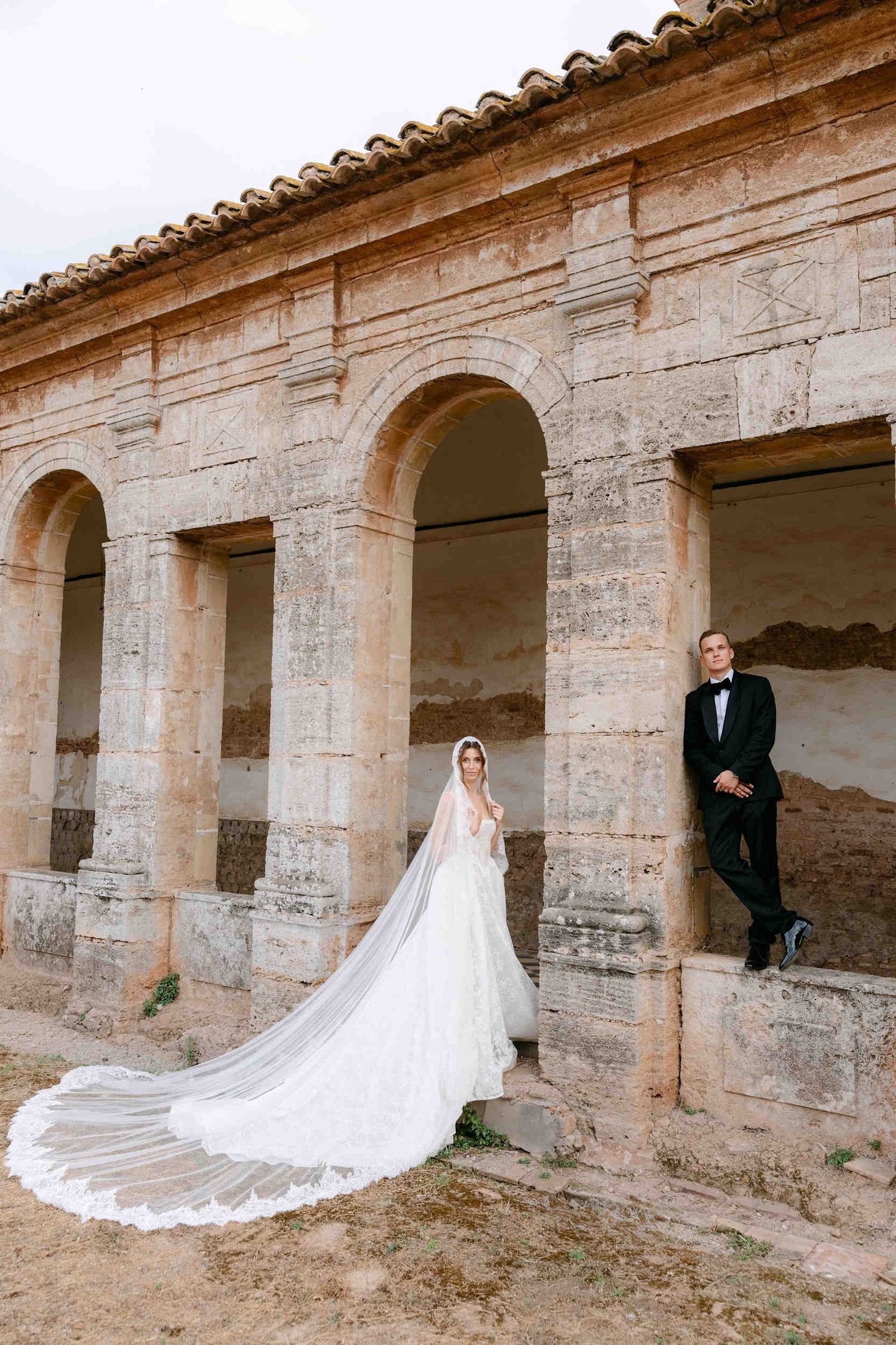 Newlyweds relaxing by the rustic stone pillars of La Cartuja de Ara Christi, with the bride's lace train elegantly spread.