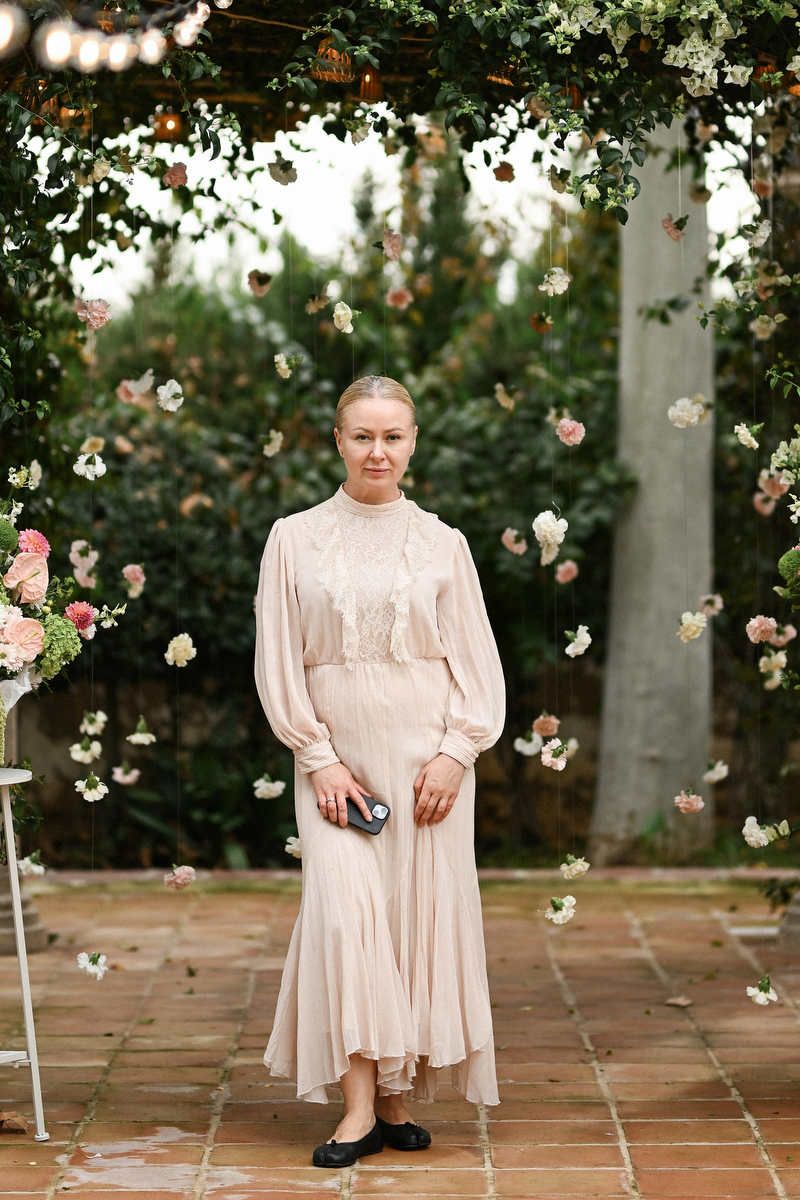 Wedding planner in Valencia Olga Prudko, stands under a garden floral archway in a beige dress holding her phone.