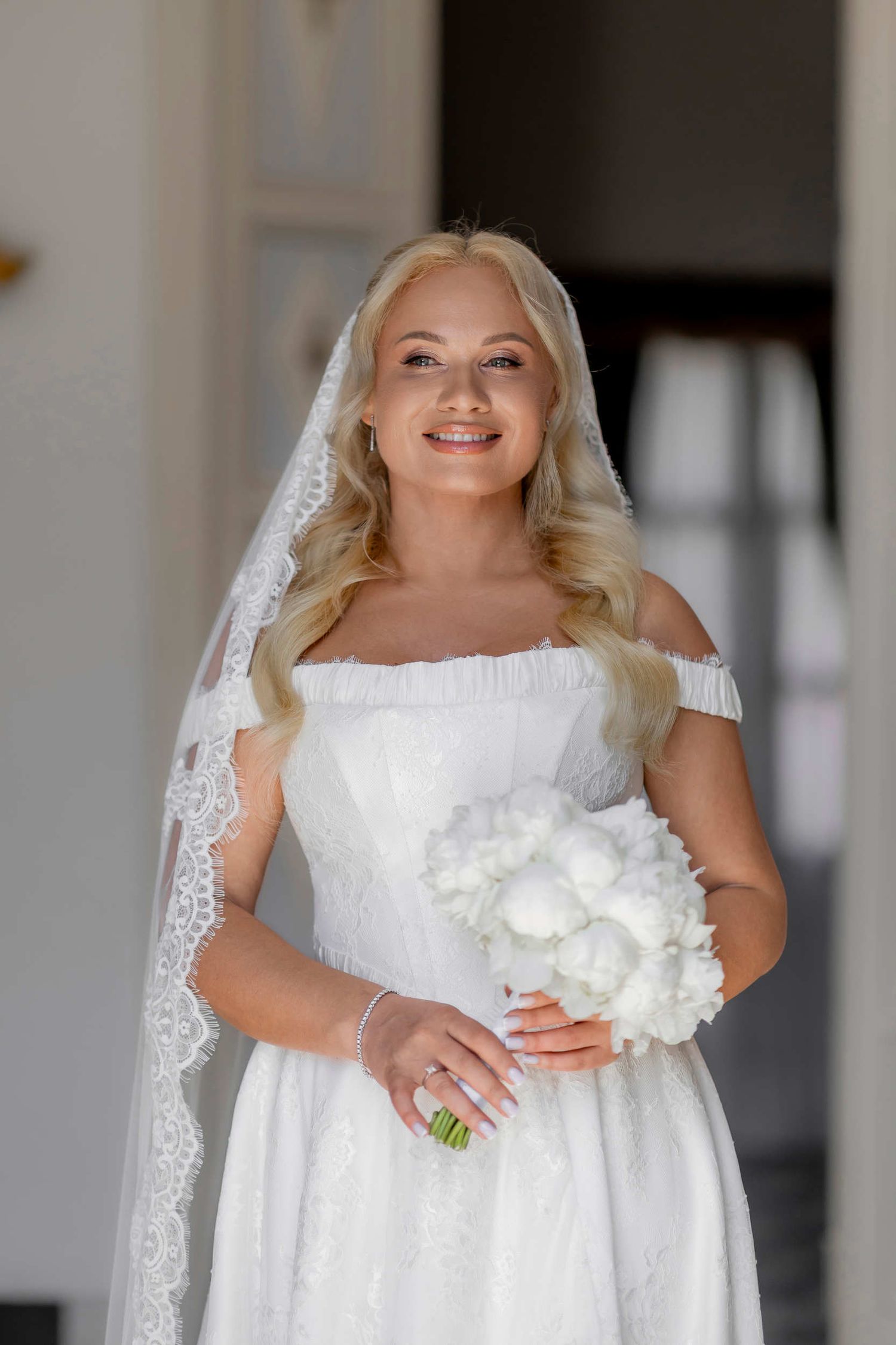 Smiling bride in a white lace dress with a veil holding a white bouquet before her Finca Marques de Montemolar wedding in Altea.