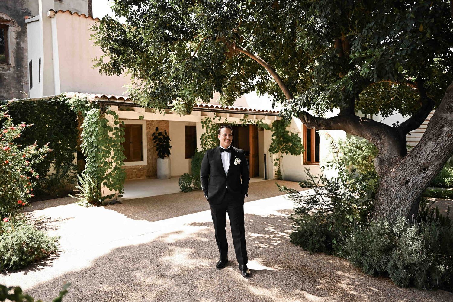 Groom in a black tuxedo smiling outside under a large tree in the gardens of Finca del Canónigo in Valencia.