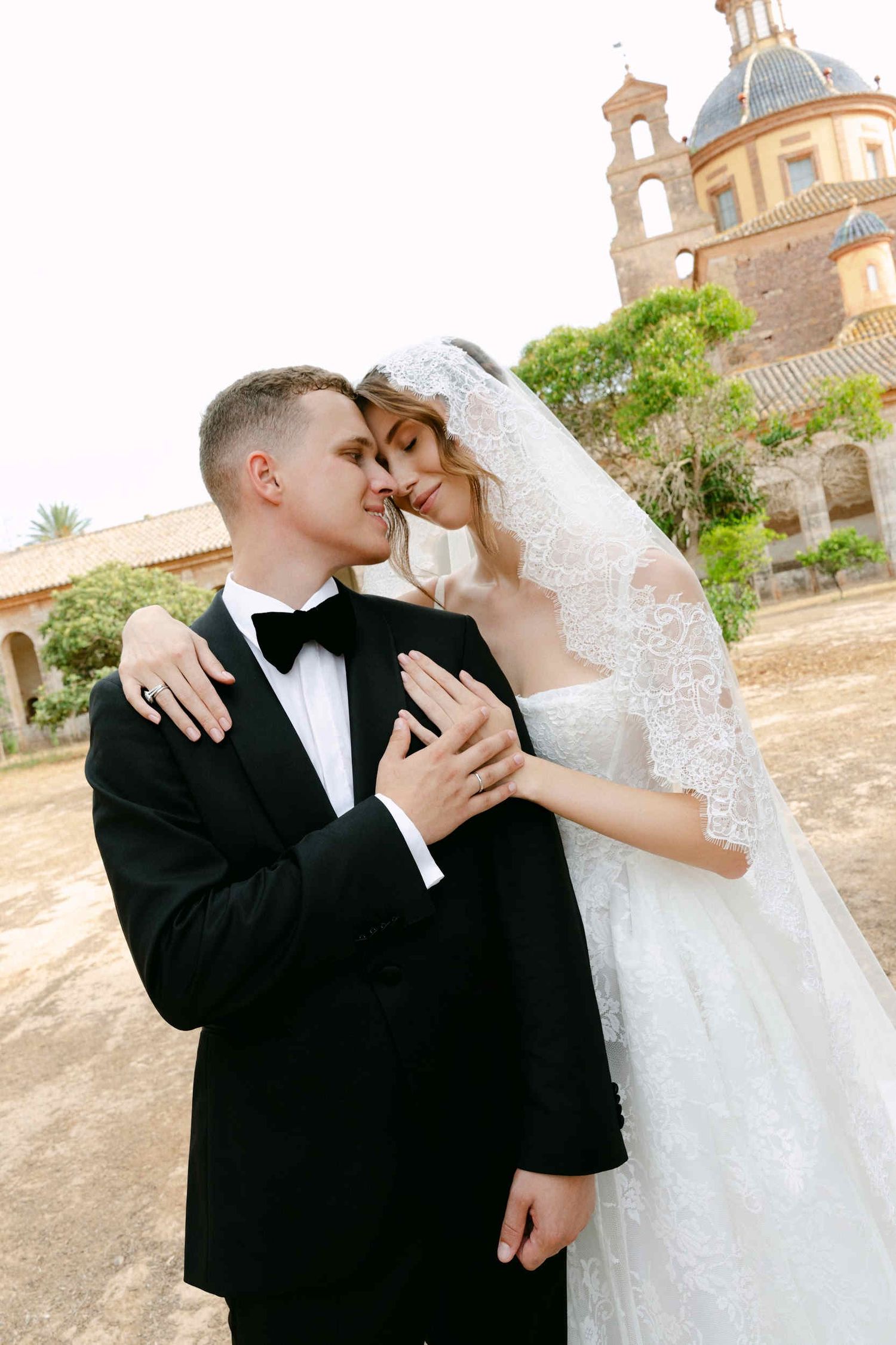 Groom in a tuxedo holding his bride who wears a traditional lace mantilla veil, posing affectionately at a monastery cartuja ara christi