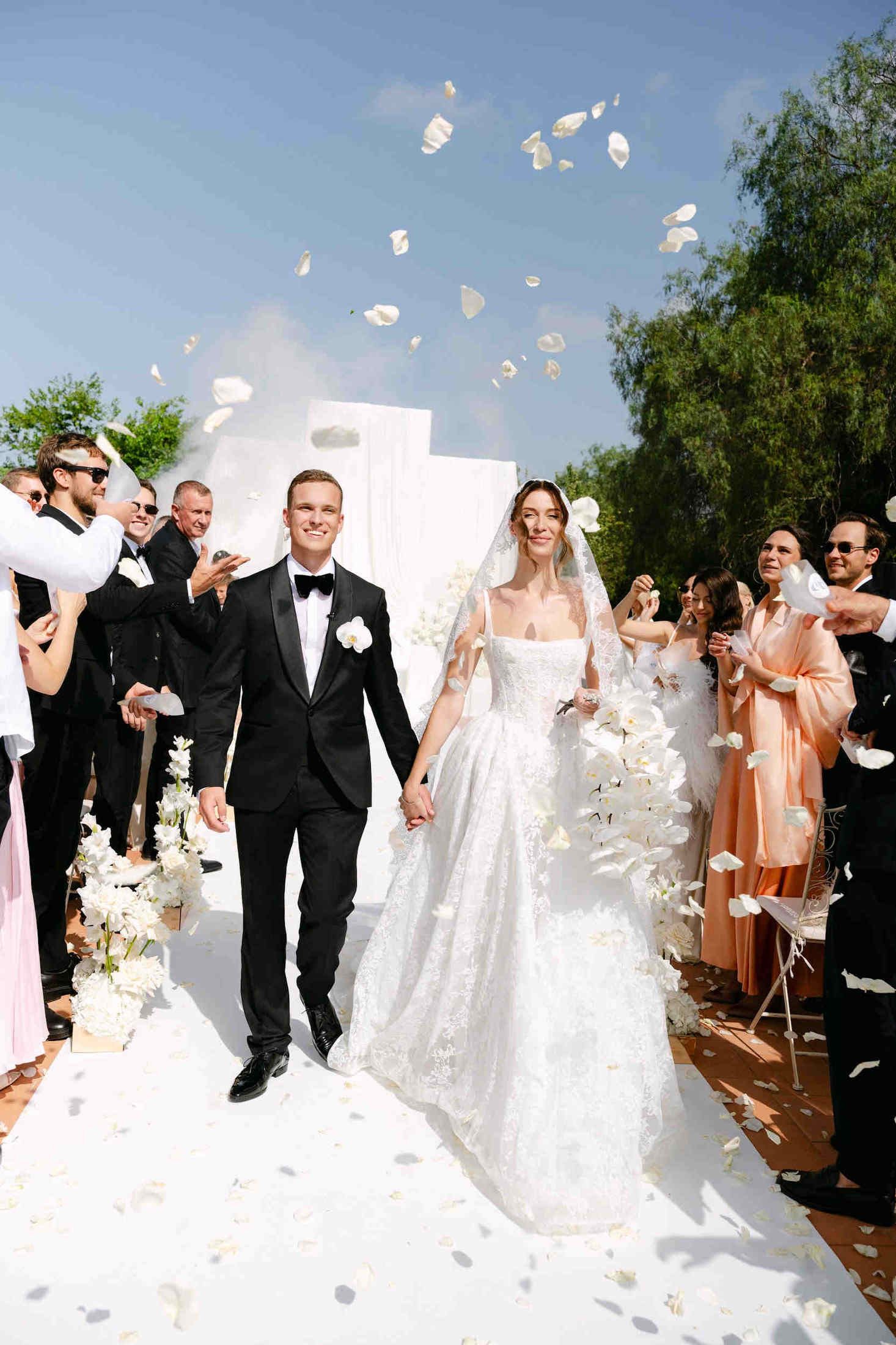 Newlyweds holding hands and smiling as guests throw white petals during their bright outdoor wedding exit in Valencia.