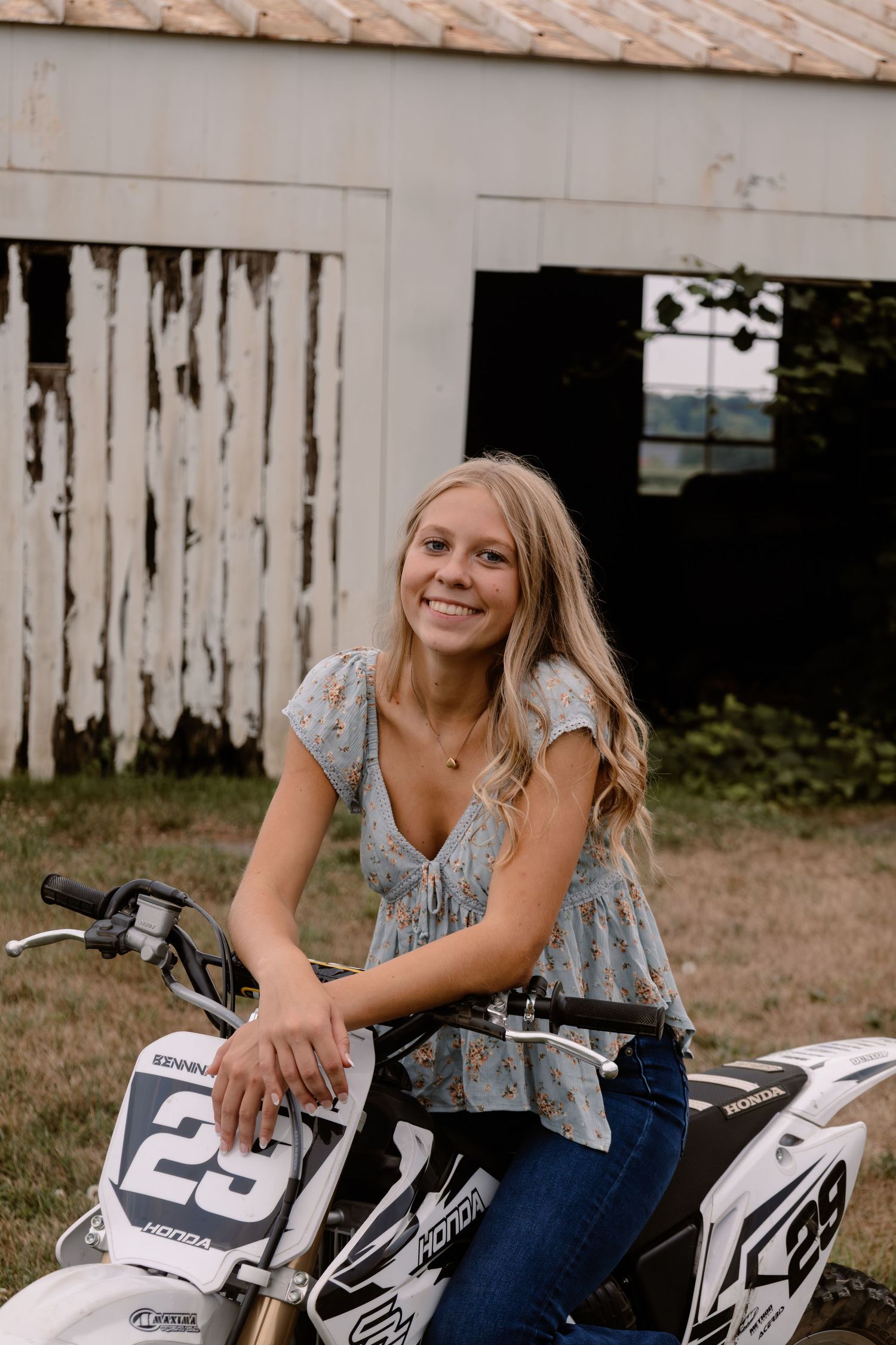 Young blonde woman smiling while sitting on a white Honda dirt bike numbered 29 outdoors near a rustic barn.