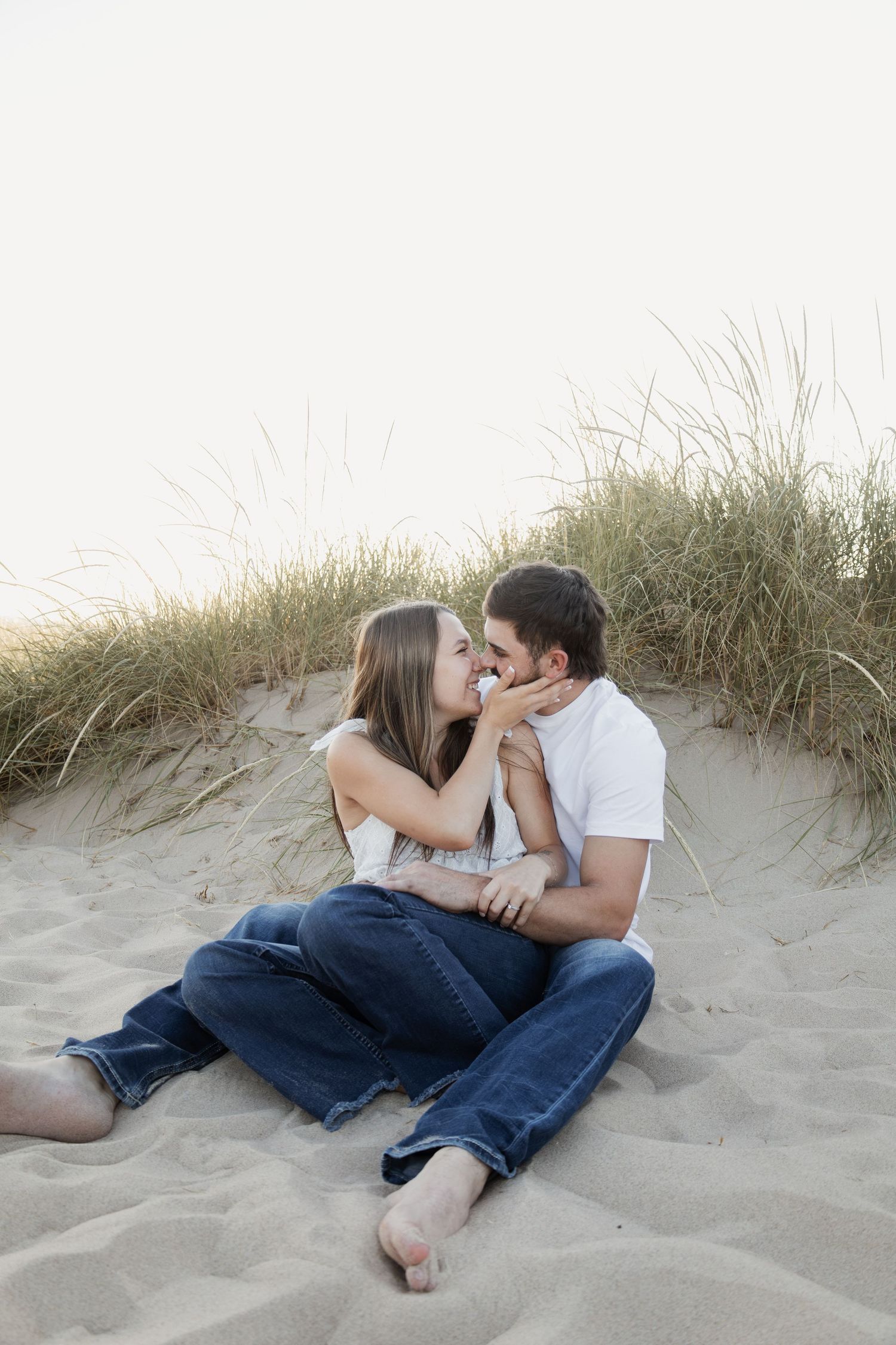 Romantic couple sitting on sandy beach dunes, sharing a tender moment, wearing jeans and casual tops.