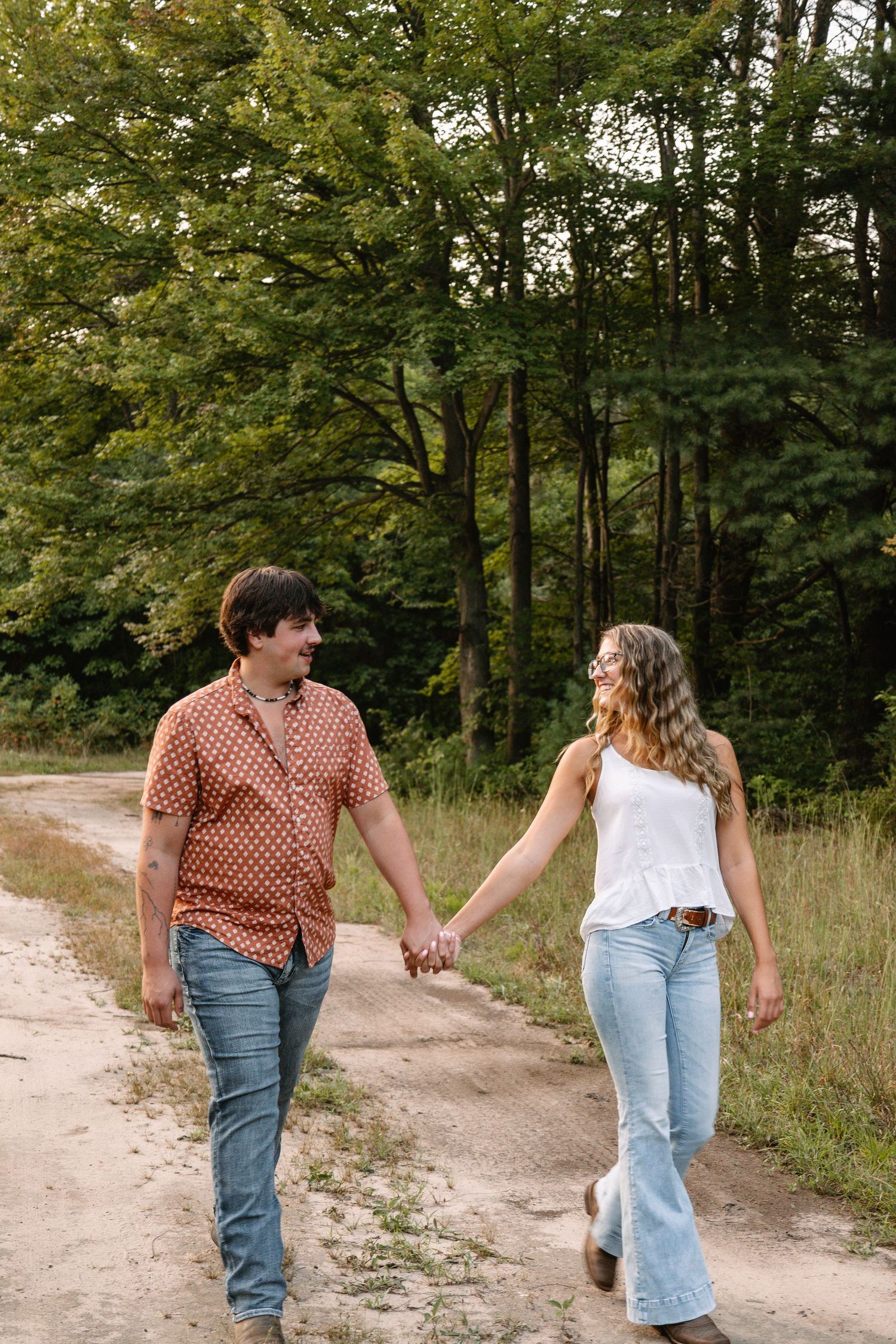 Couple holding hands and smiling at each other while walking on a dirt path surrounded by lush green trees.